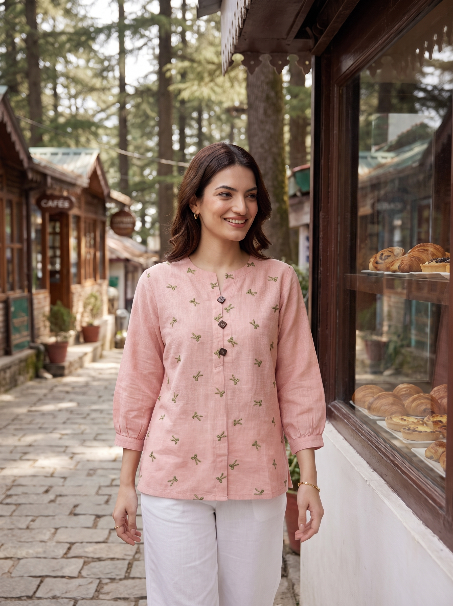 A woman stands on a cobblestone street in front of a bakery shop, wearing a light pink linen kurta with small green floral prints and white pants.
