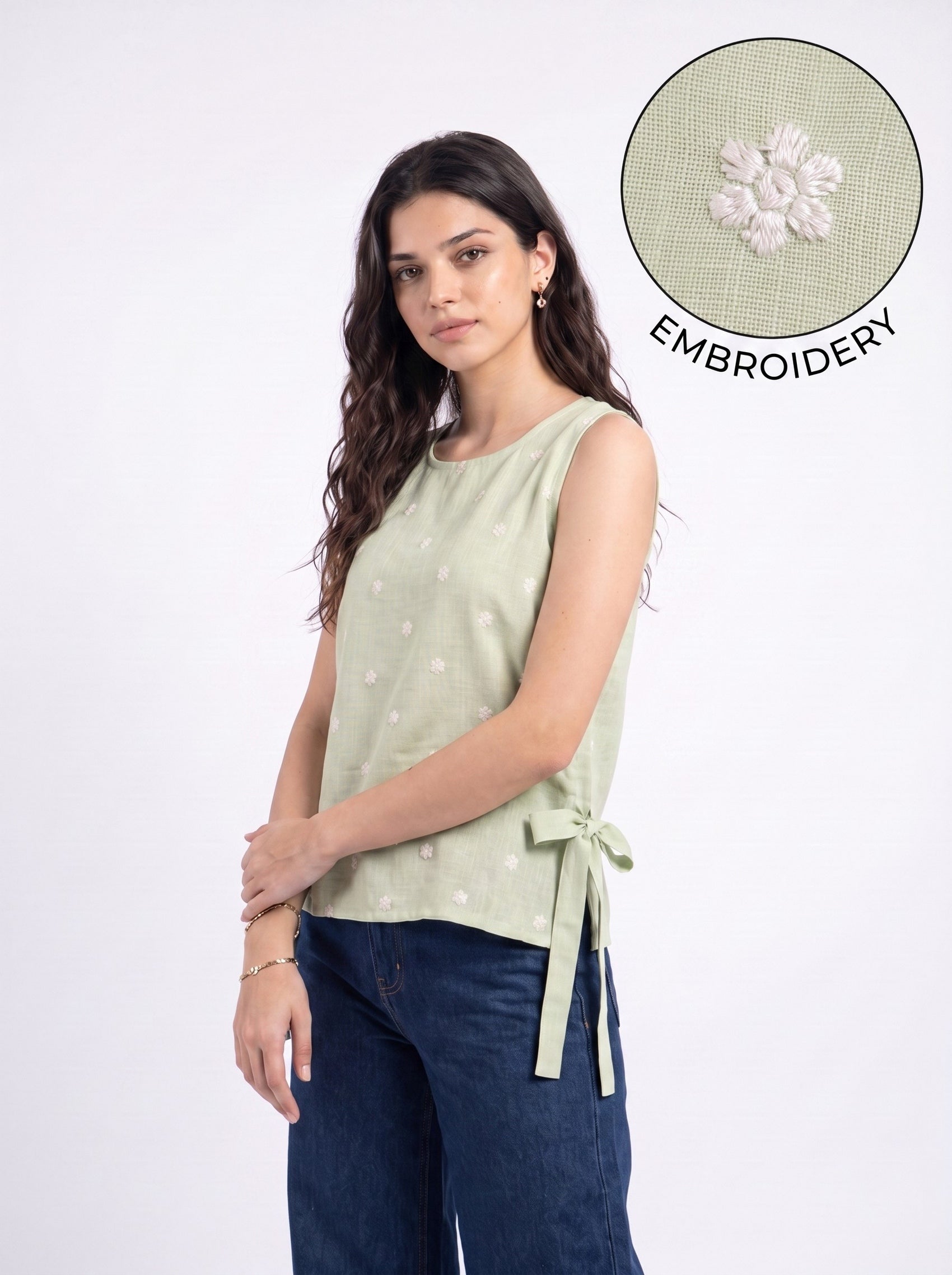 A woman modeling a sleeveless sage green top with small embroidered details and a side tie, paired with dark blue jeans. A circular inset in the top right shows a close-up of the fabric embroidery.