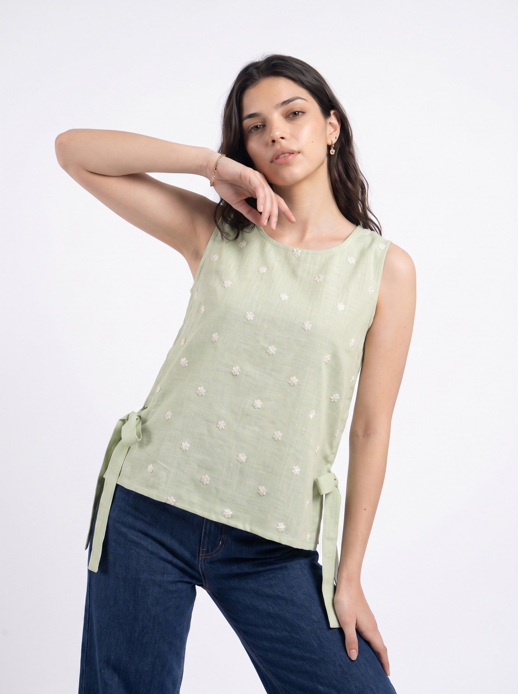 A woman modeling a sleeveless sage green top with white embroidered floral details and side ties, paired with dark blue jeans, against a plain white studio background.