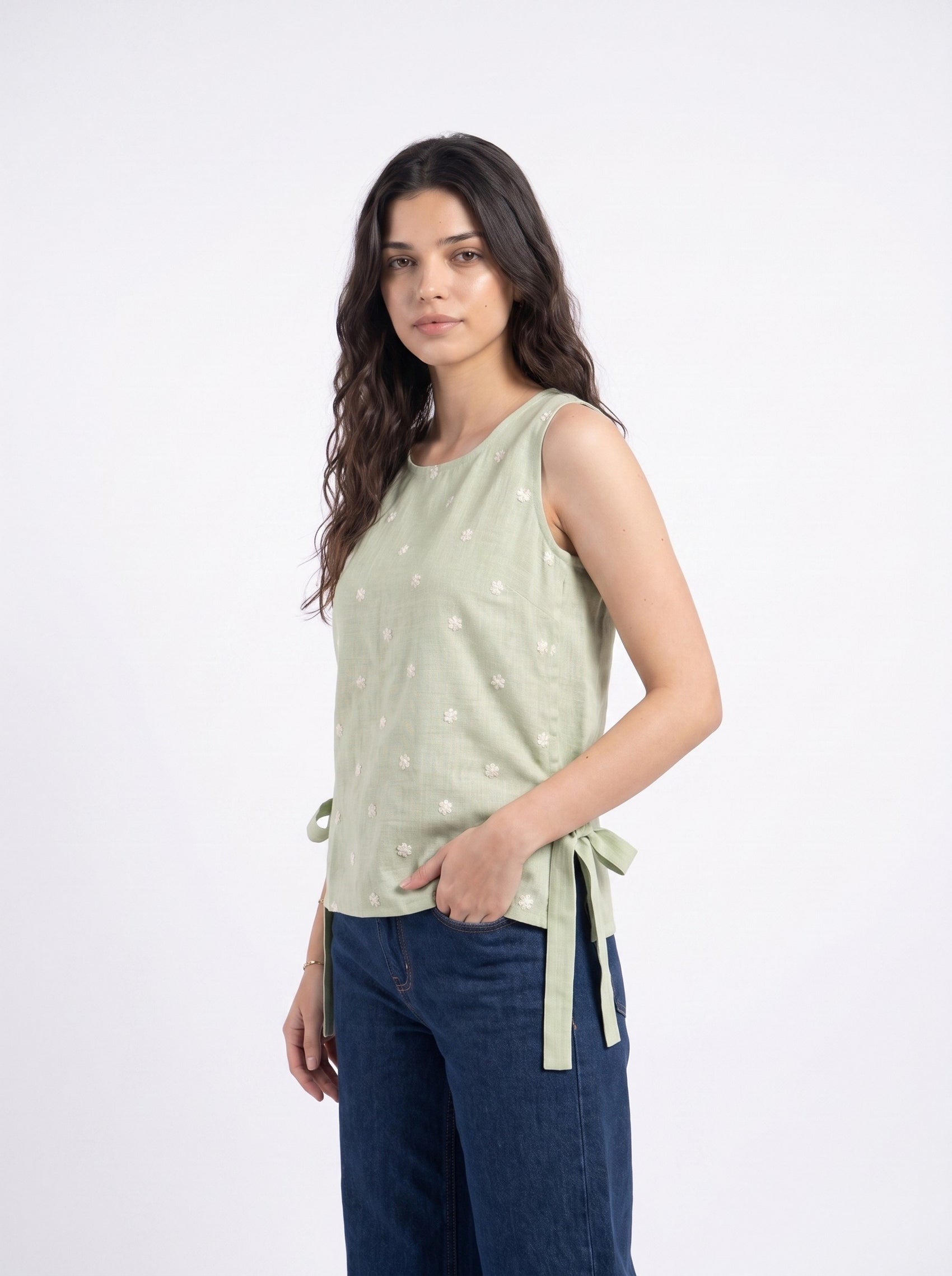 A woman modeling a sleeveless sage green top with small white embroidered details and side tie details, paired with dark blue jeans, against a plain white studio background.