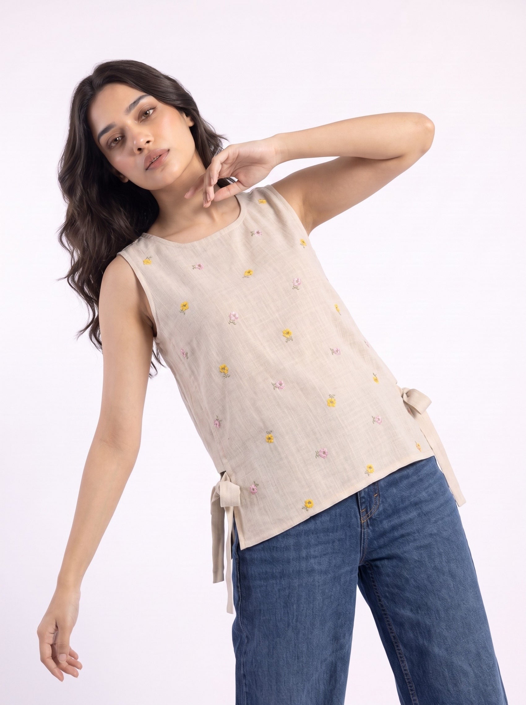 A woman modeling a sleeveless beige linen top with small yellow and pink embroidered flowers and side tie details, paired with blue jeans, against a plain white background.