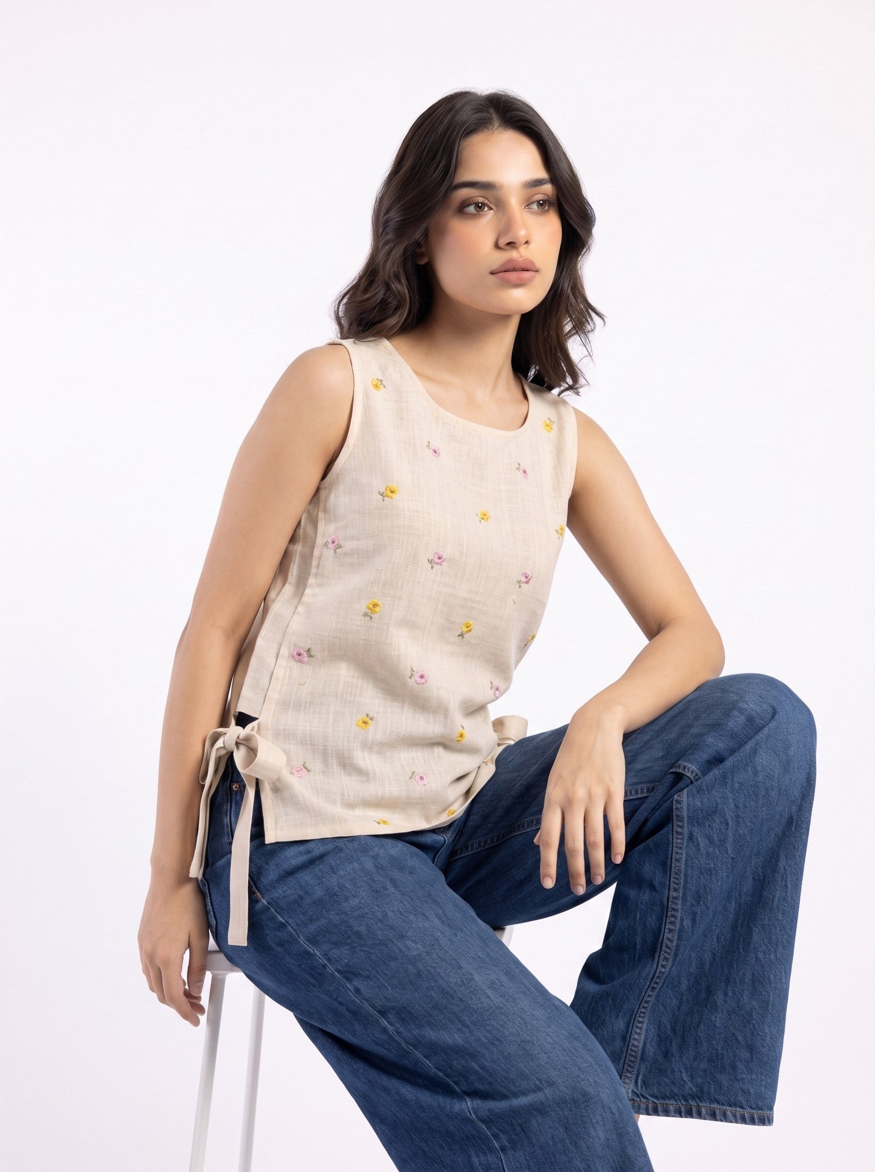 A woman wearing a sleeveless beige linen top with small yellow and pink embroidered flowers, paired with wide-leg blue jeans, sitting on a white stool against a white studio background.
