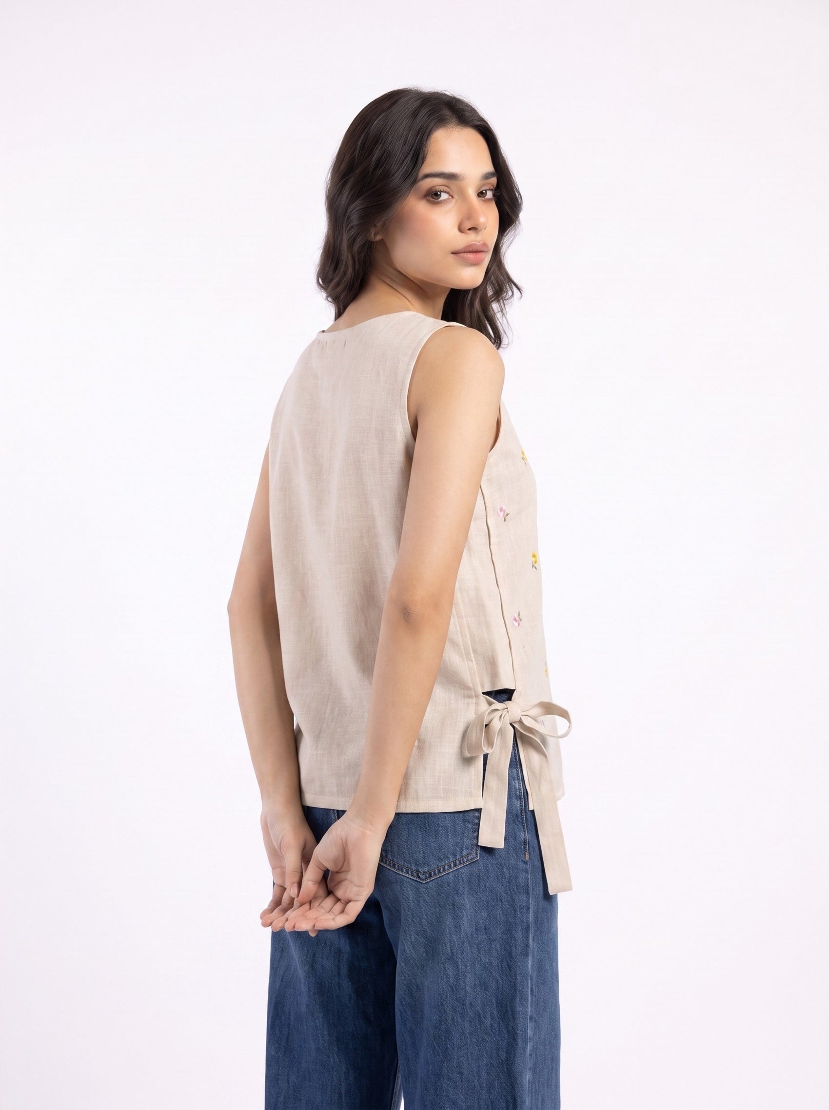 A woman wearing a sleeveless beige linen vest with a side tie detail and blue jeans, photographed from the back against a plain white studio background.