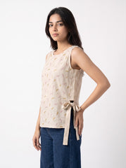 A woman modeling a sleeveless beige top with a subtle gold and white pattern, featuring a side tie detail, paired with dark blue jeans against a plain white background.