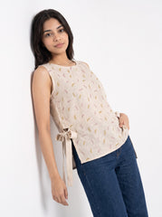A woman wearing a sleeveless beige top with a delicate gold and green leaf pattern, paired with dark blue jeans, posing against a plain white wall.