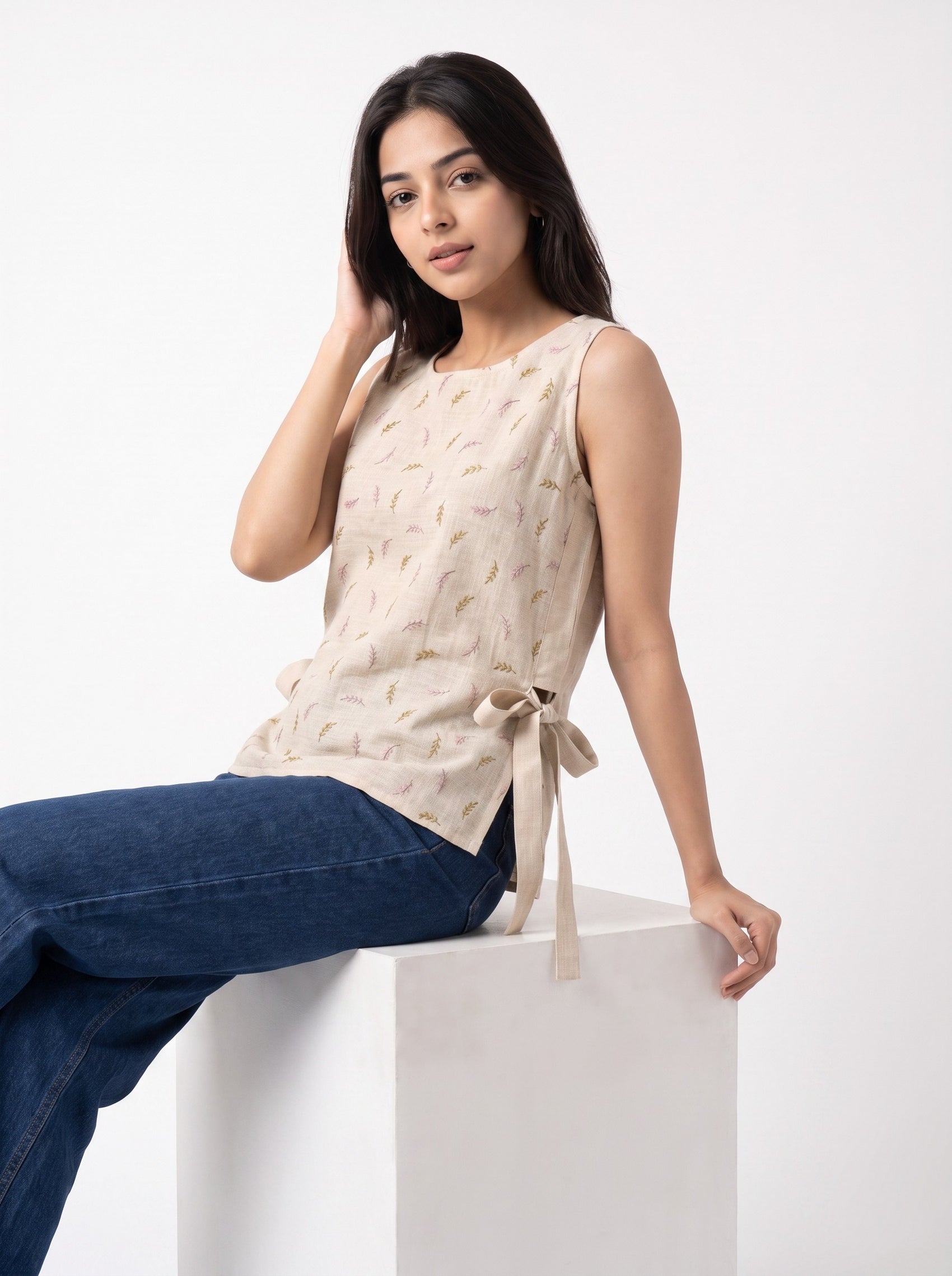A woman wearing a sleeveless beige top with a subtle leaf pattern and dark blue jeans, sitting on a white block against a white studio background.
