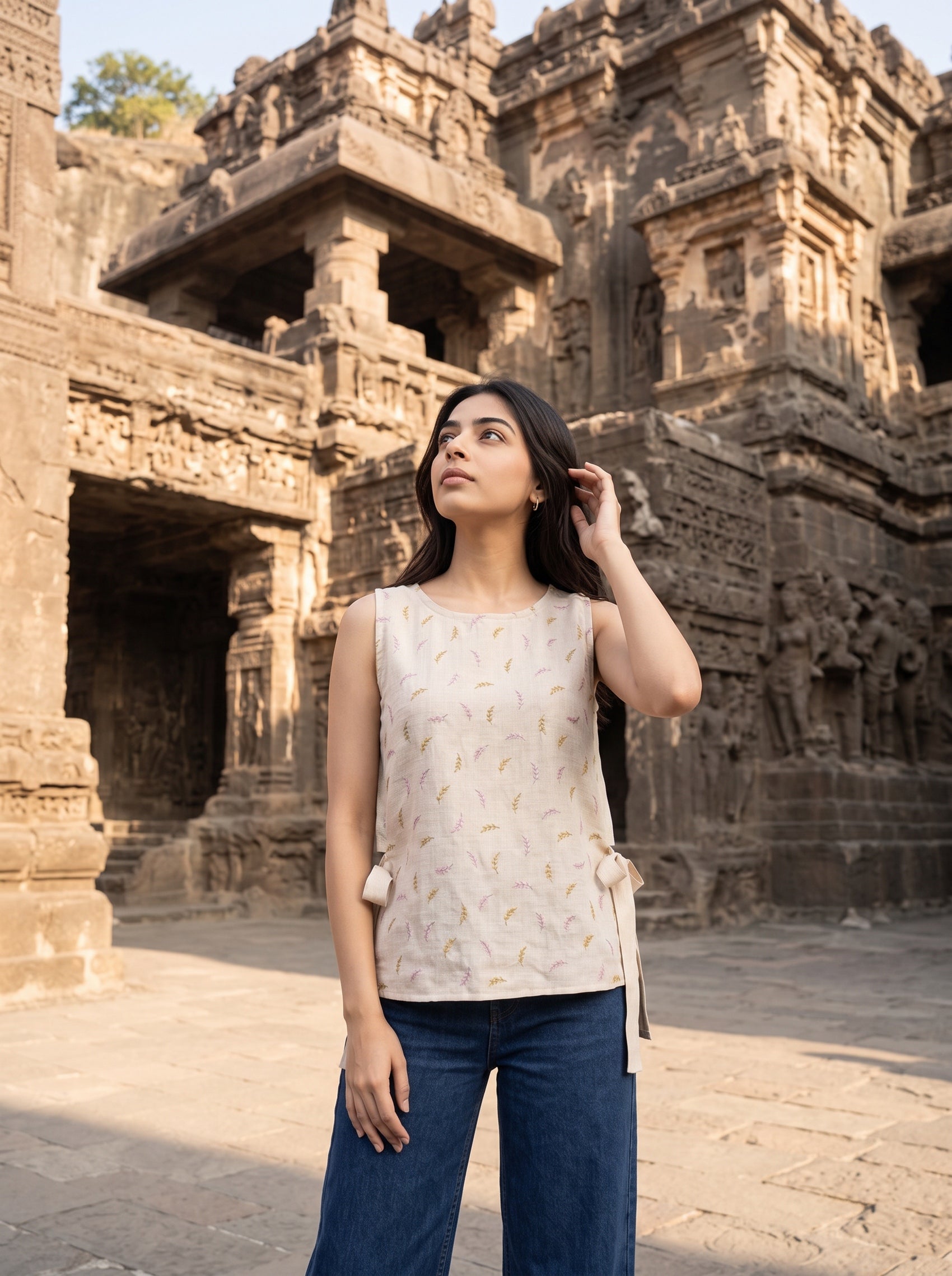 A woman wearing a sleeveless cream top with a subtle gold leaf pattern and blue jeans stands in front of an ancient stone temple.
