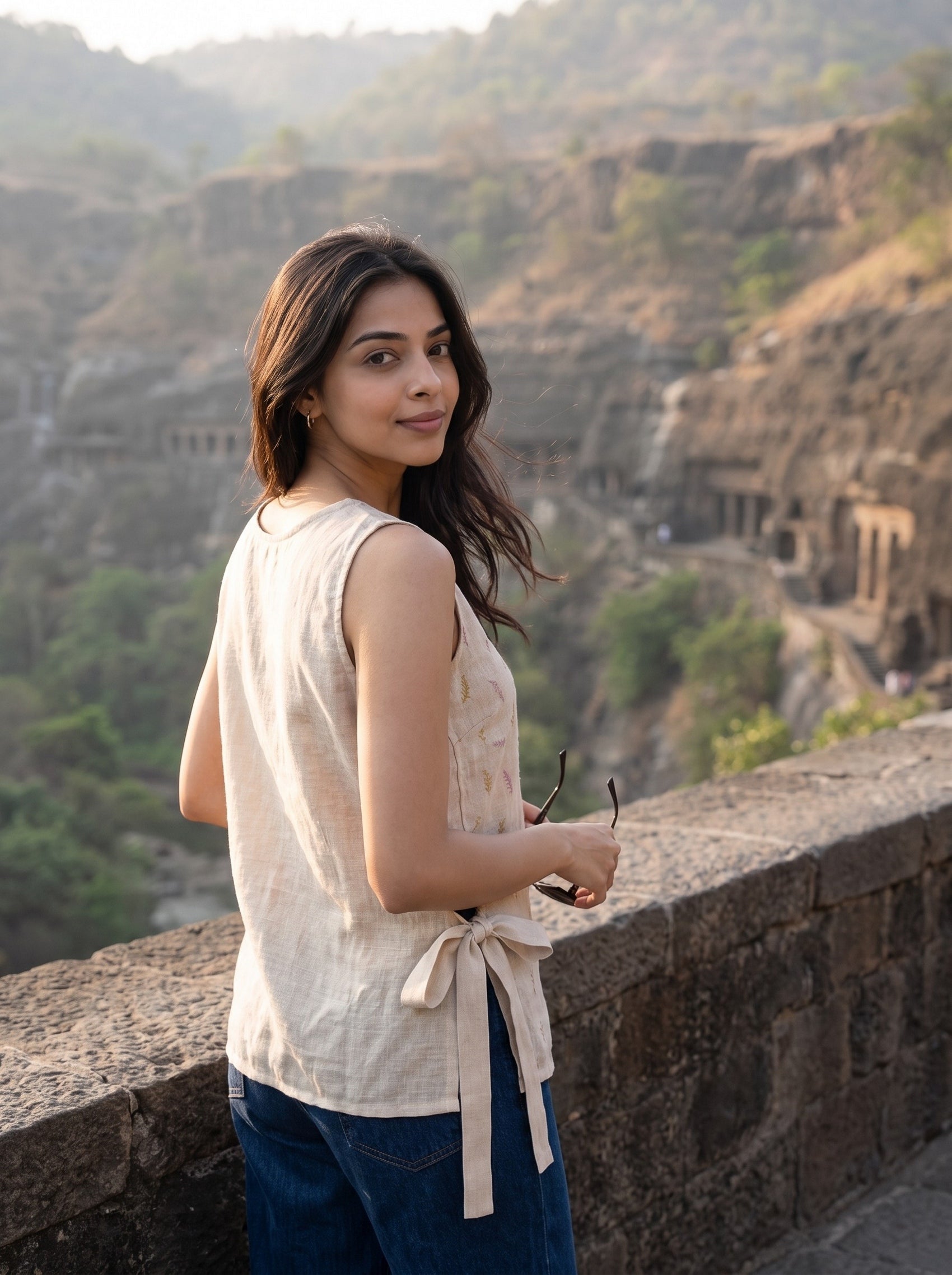 A woman wearing a sleeveless cream linen top and blue jeans stands on a stone wall overlooking a rocky canyon with cave-like structures in the background.