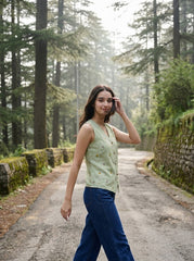 A woman walking on a forest path wearing a light sage green sleeveless top with small yellow floral prints and dark blue wide-leg jeans.