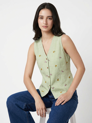 A woman seated on a white stool wearing a sleeveless light sage green vest with small embroidered floral details and blue jeans, photographed against a clean white studio background.