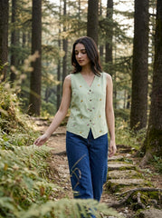 A woman walking on a forest path wearing a sleeveless light green vest with small embroidered details and blue jeans.