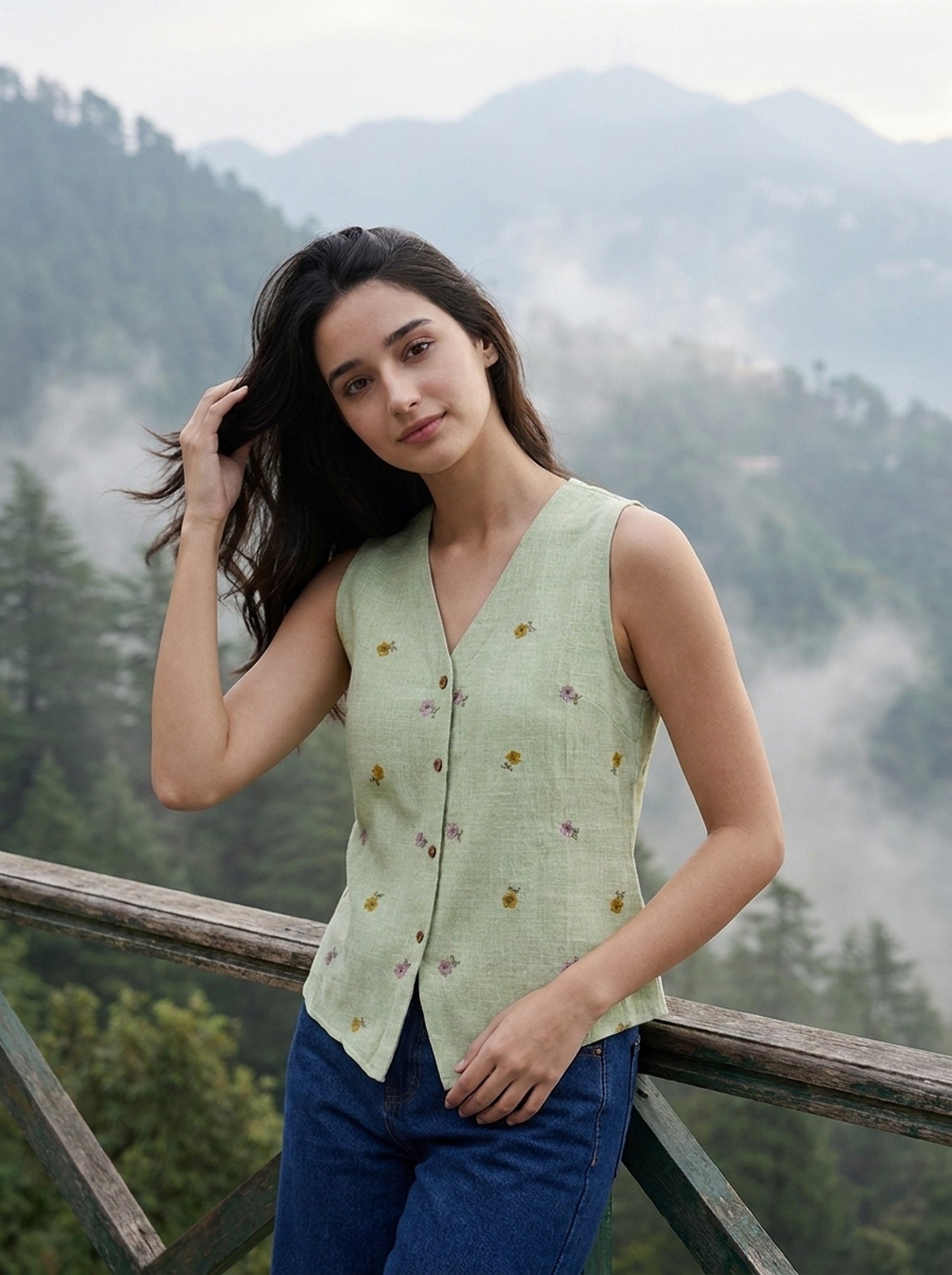 A woman wearing a light sage green sleeveless vest with small yellow and purple floral embroidery, paired with blue jeans, posing outdoors against a misty mountain forest background.