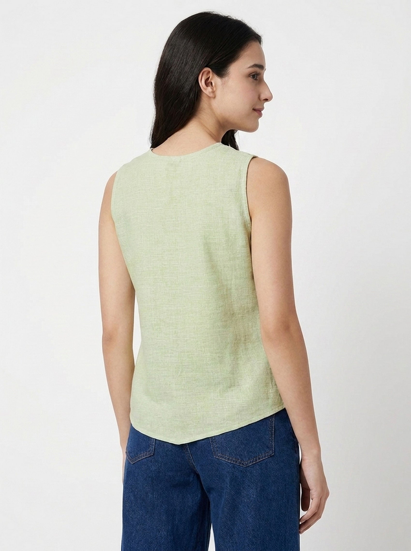 A woman shown from the back wearing a light sage green sleeveless top and blue jeans, photographed against a plain light gray studio background.