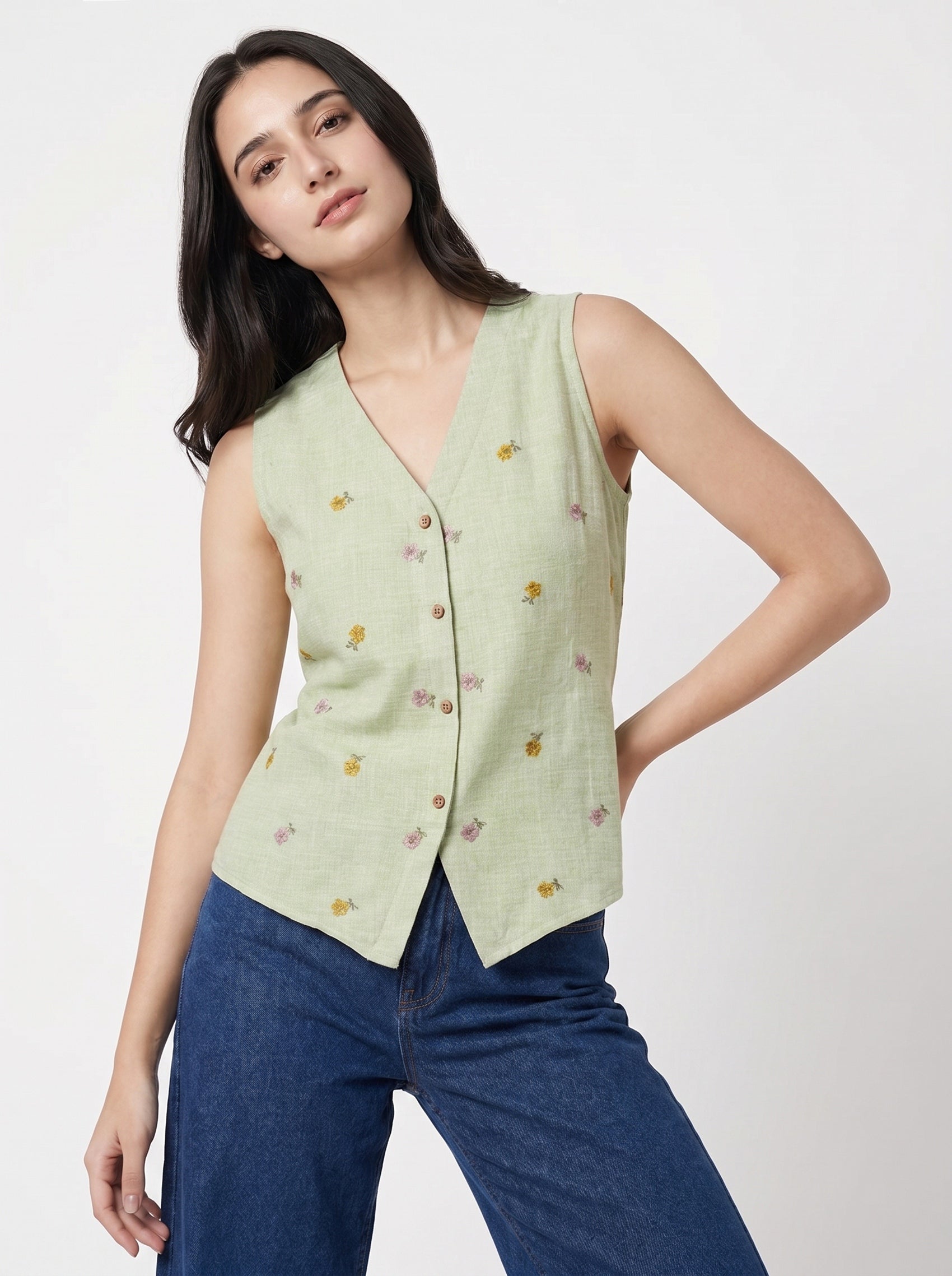 A woman modeling a sleeveless light sage green vest with a V-neck, button front, and small embroidered floral details, paired with blue jeans against a plain light gray studio background.