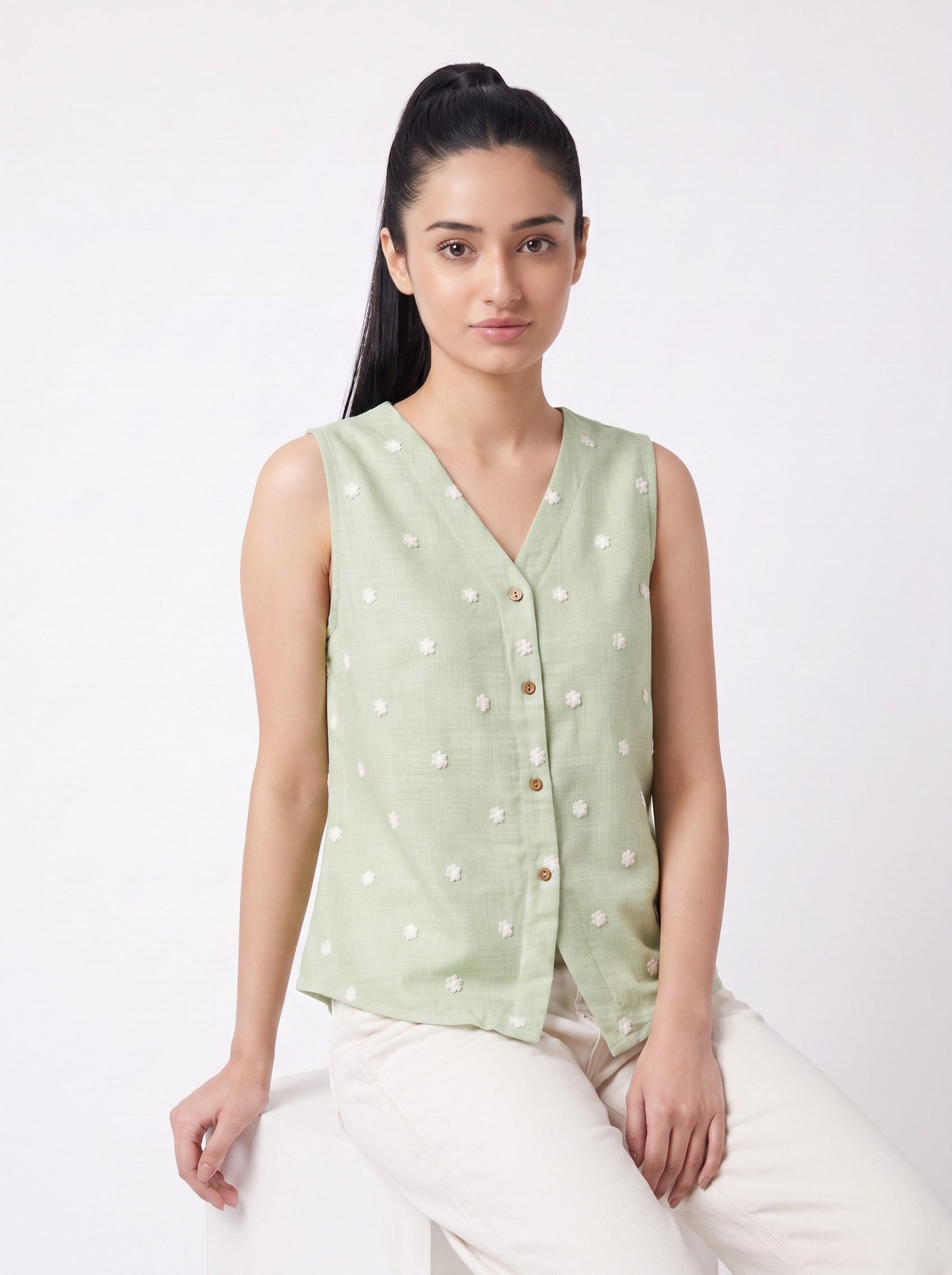 A woman wearing a sleeveless light sage green linen vest with white embroidered dots and a button-up front, paired with off-white trousers, seated against a plain white studio background.