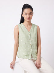 A woman wearing a sleeveless light sage green linen vest with white embroidered dots and a button-up front, paired with off-white trousers, seated against a plain white studio background.
