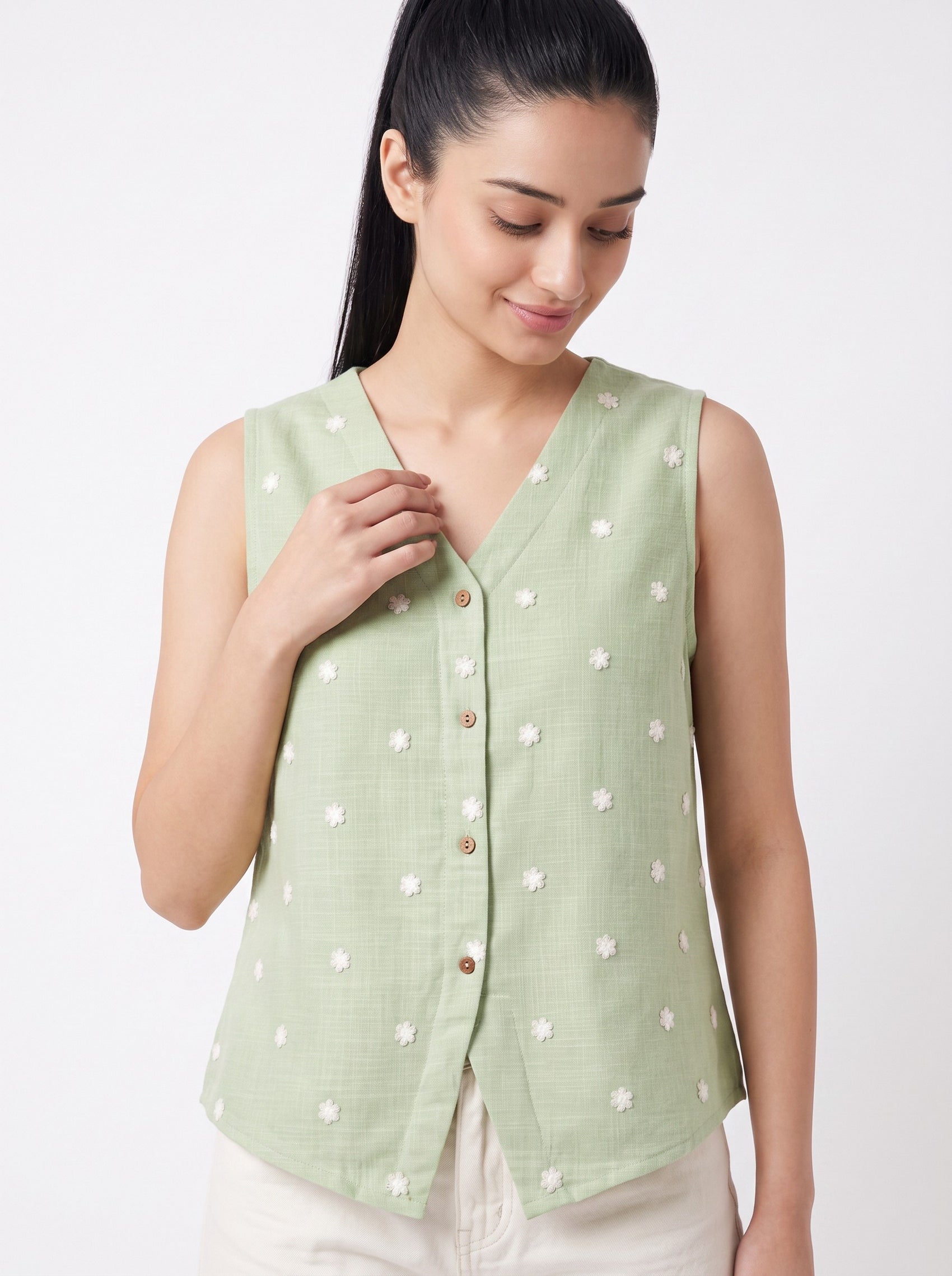 A woman wearing a sleeveless light sage green vest with white embroidered floral details and brown buttons, paired with off-white pants, against a plain white studio background.