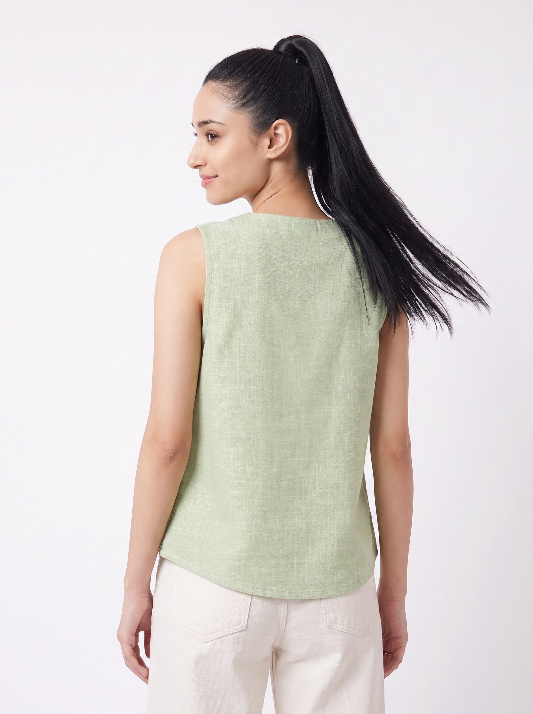 A woman with long black hair in a ponytail is shown from the back wearing a light sage green sleeveless top and cream pants against a plain white studio background.