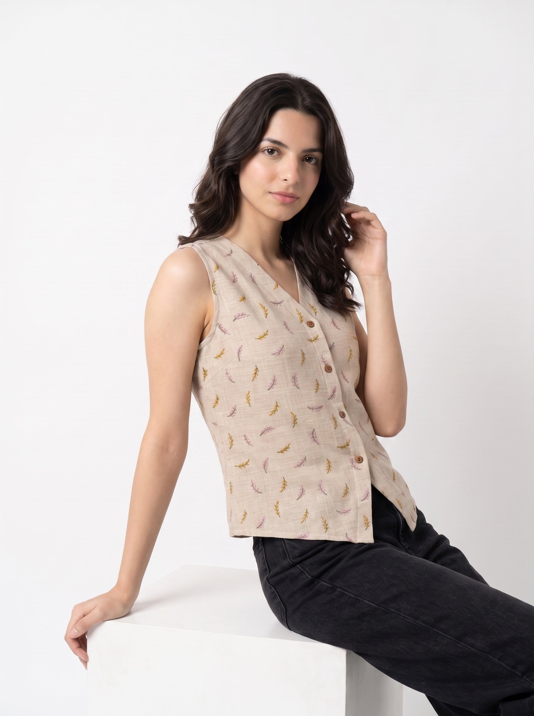 A woman wearing a sleeveless beige vest with a small multicolor feather pattern and dark jeans, posing on a white block against a white studio background.