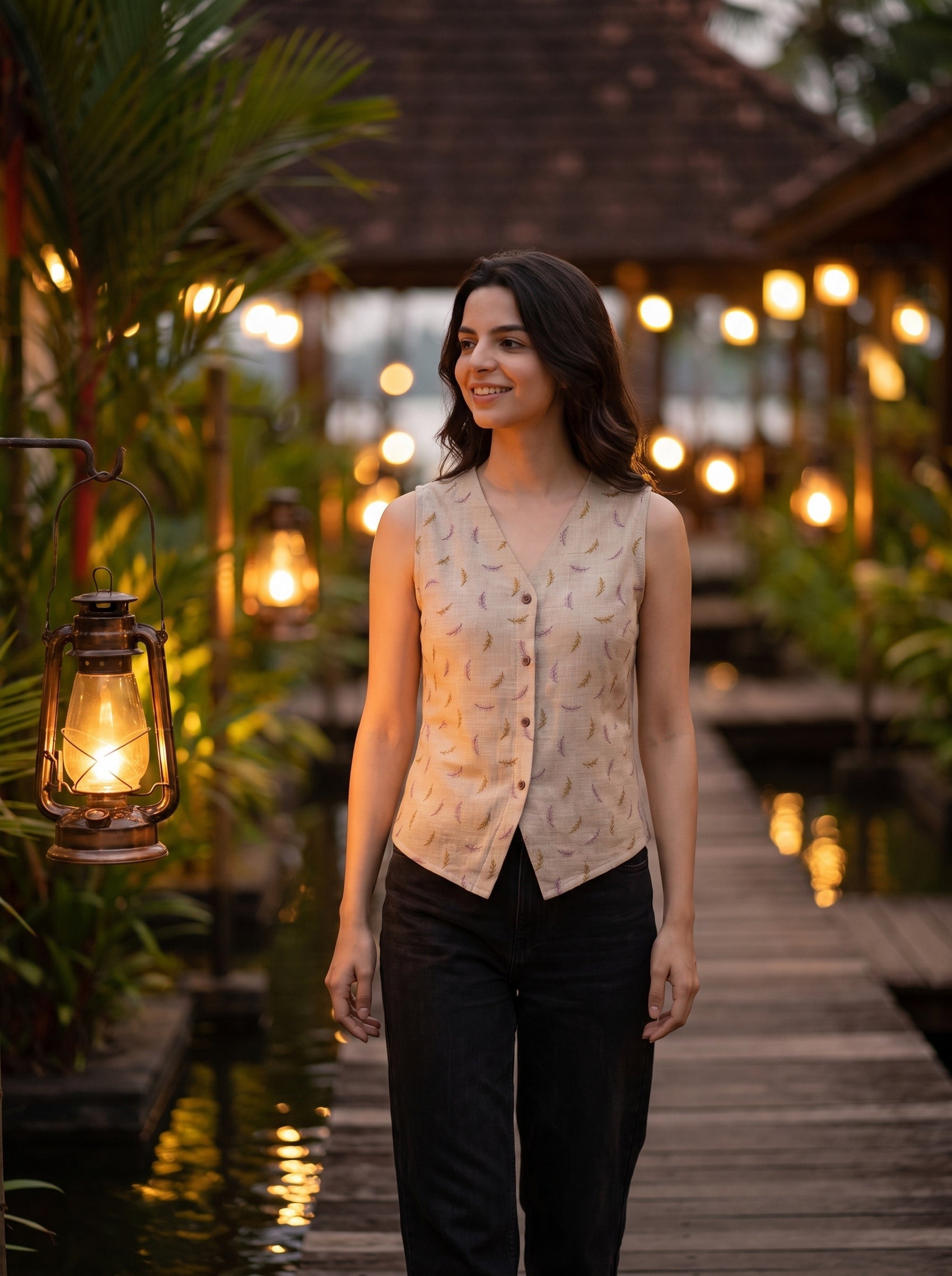 A woman stands on a wooden walkway at dusk wearing a sleeveless beige vest with a subtle leaf pattern and black pants. Warm lantern lights and tropical foliage are visible in the background.