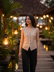 A woman stands on a wooden walkway at dusk wearing a sleeveless beige vest with a subtle leaf pattern and black pants. Warm lantern lights and tropical foliage are visible in the background.