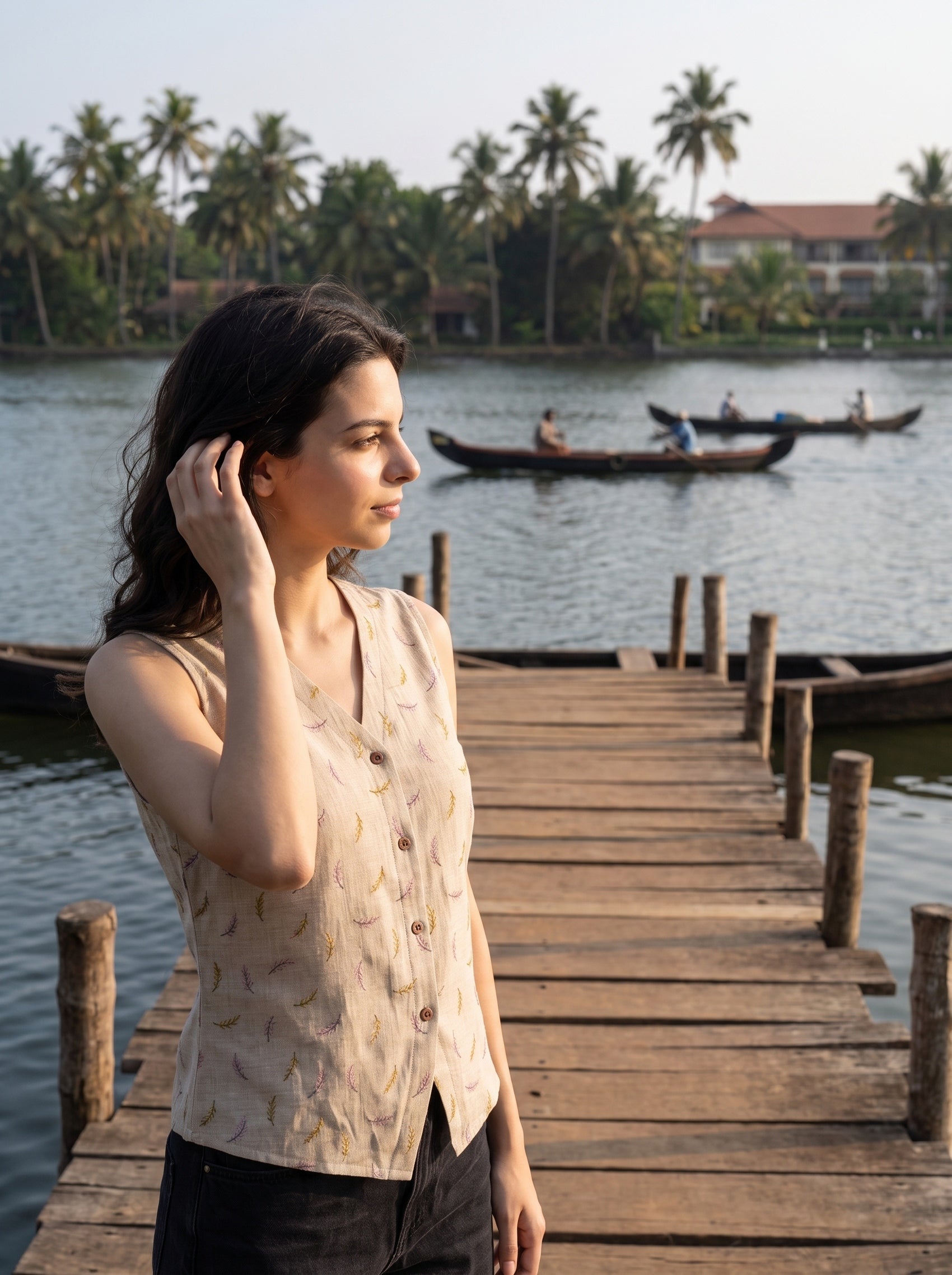 A woman stands on a wooden dock by a lake, wearing a sleeveless beige top with a subtle leaf pattern. Palm trees, water, and small boats are visible in the background.