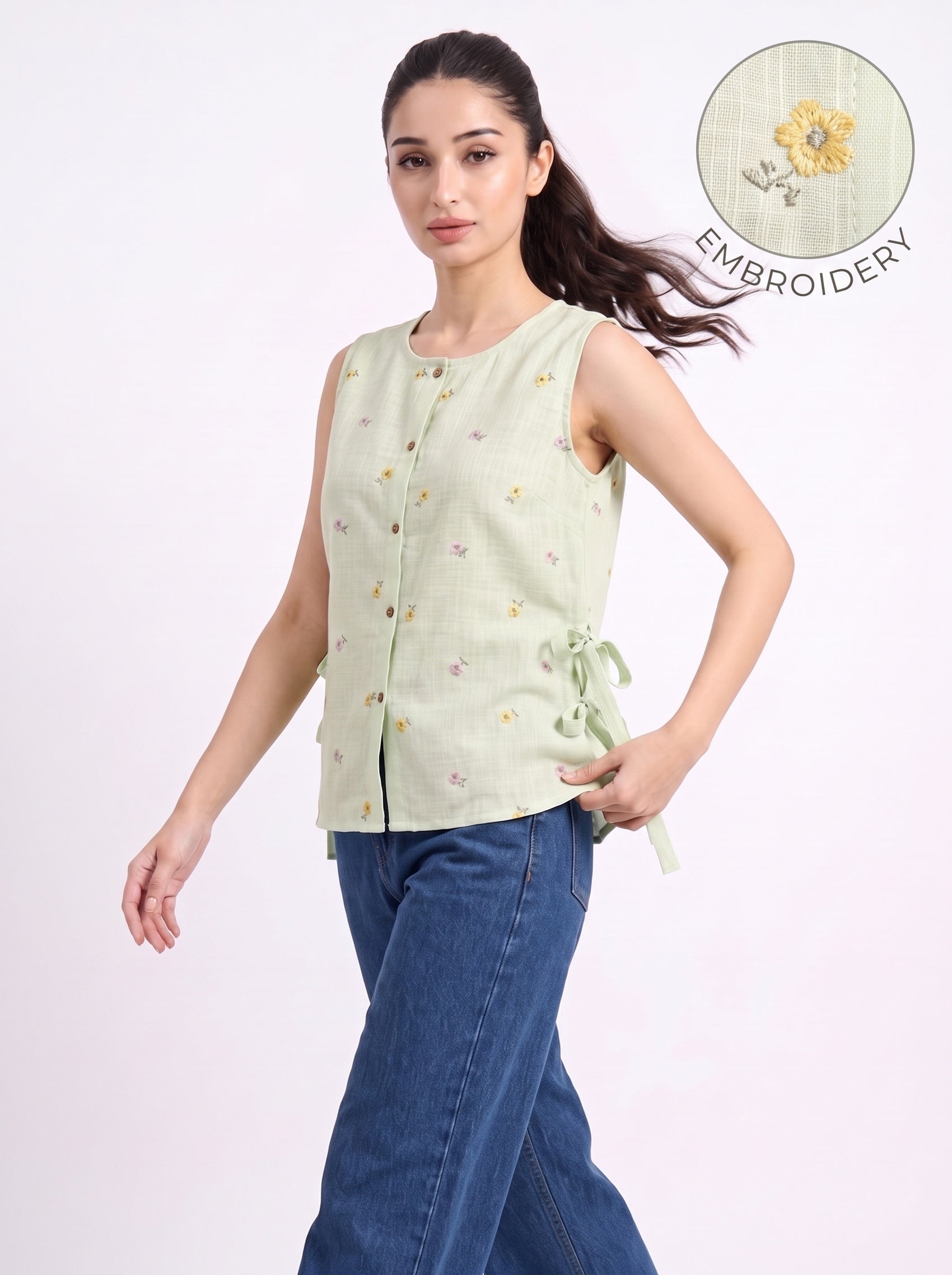 A woman modeling a sleeveless light green top with small embroidered floral details and side ties, paired with blue jeans, against a plain light background. An inset circle in the top right shows a close-up of the embroidery.