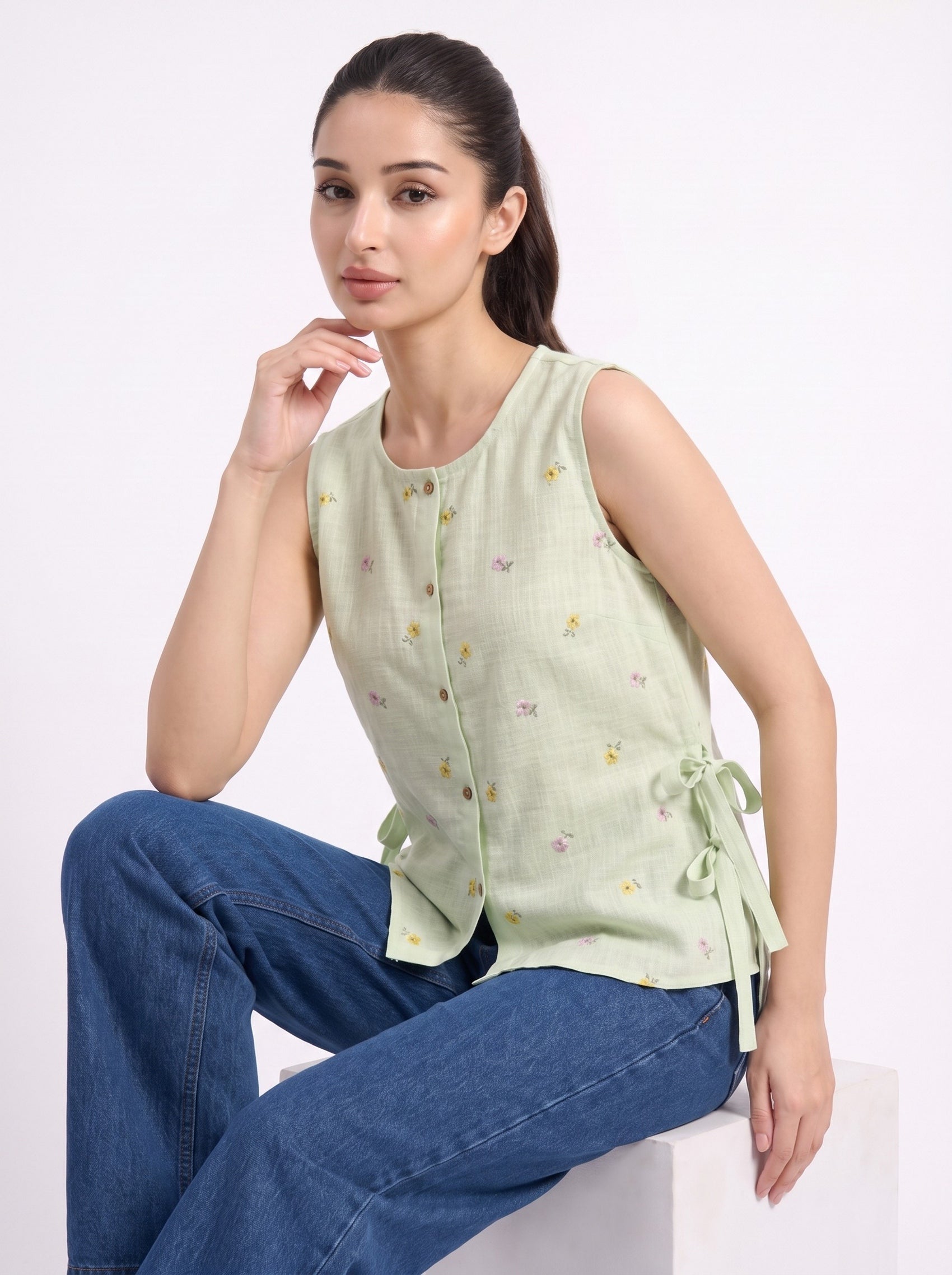 A woman wearing a light sage green sleeveless top with small embroidered motifs and side ties, paired with blue jeans, seated against a plain white background.