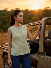A woman stands beside an open safari-style vehicle at sunset, wearing a sleeveless light green button-front top with small embroidered floral details and blue jeans.