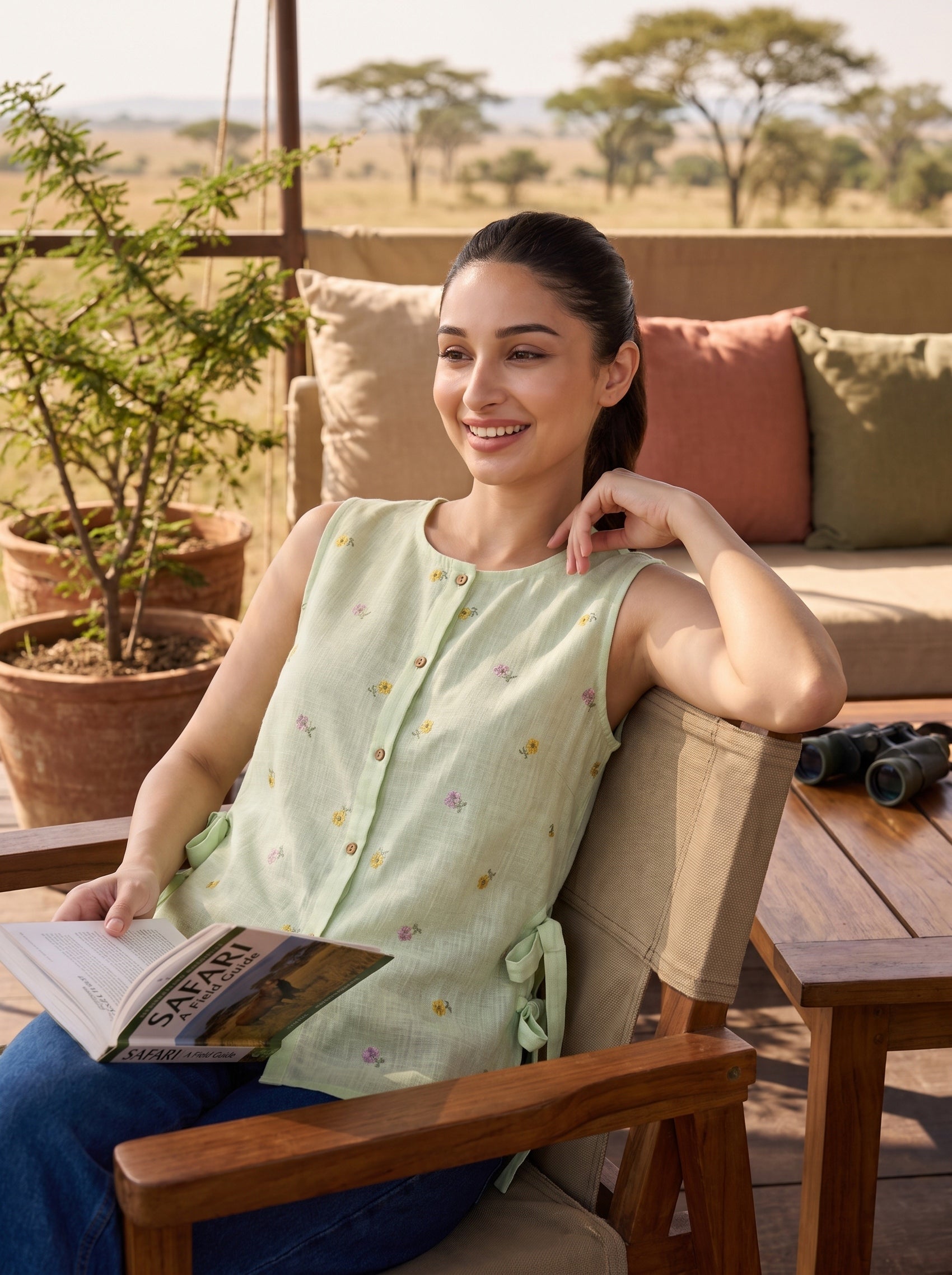 A woman sitting in a wooden lounge chair on a patio, wearing a sleeveless light green top with small embroidered motifs. She is holding a magazine, with a sofa, potted plants, and an outdoor savanna view in the background.