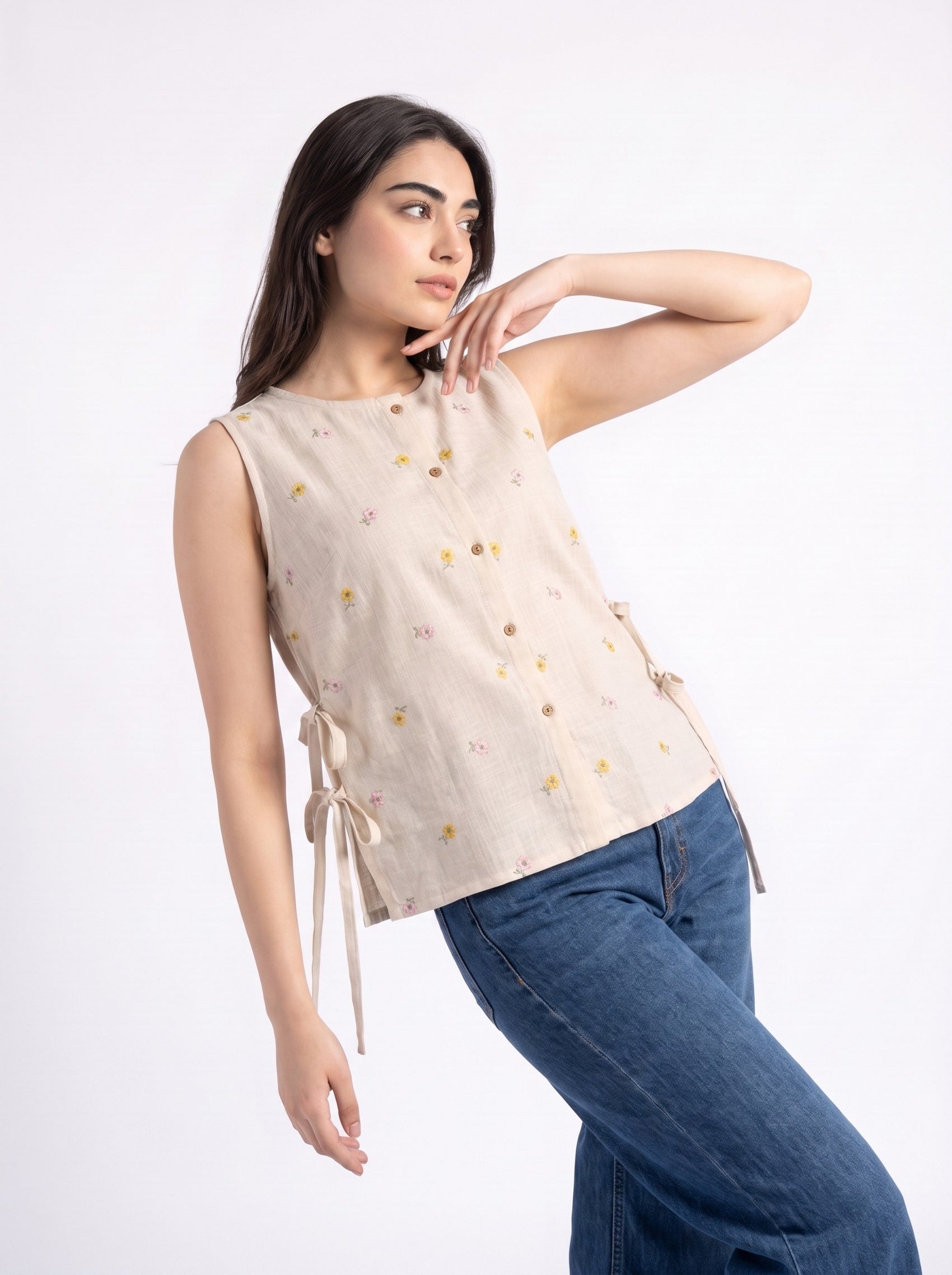 A woman modeling a sleeveless beige top with small yellow and green floral prints, side tie details, and a button front, paired with blue jeans against a plain white background.