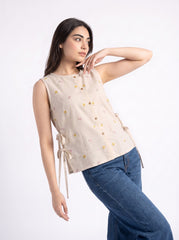 A woman modeling a sleeveless beige top with small yellow and green floral prints, side tie details, and a button front, paired with blue jeans against a plain white background.