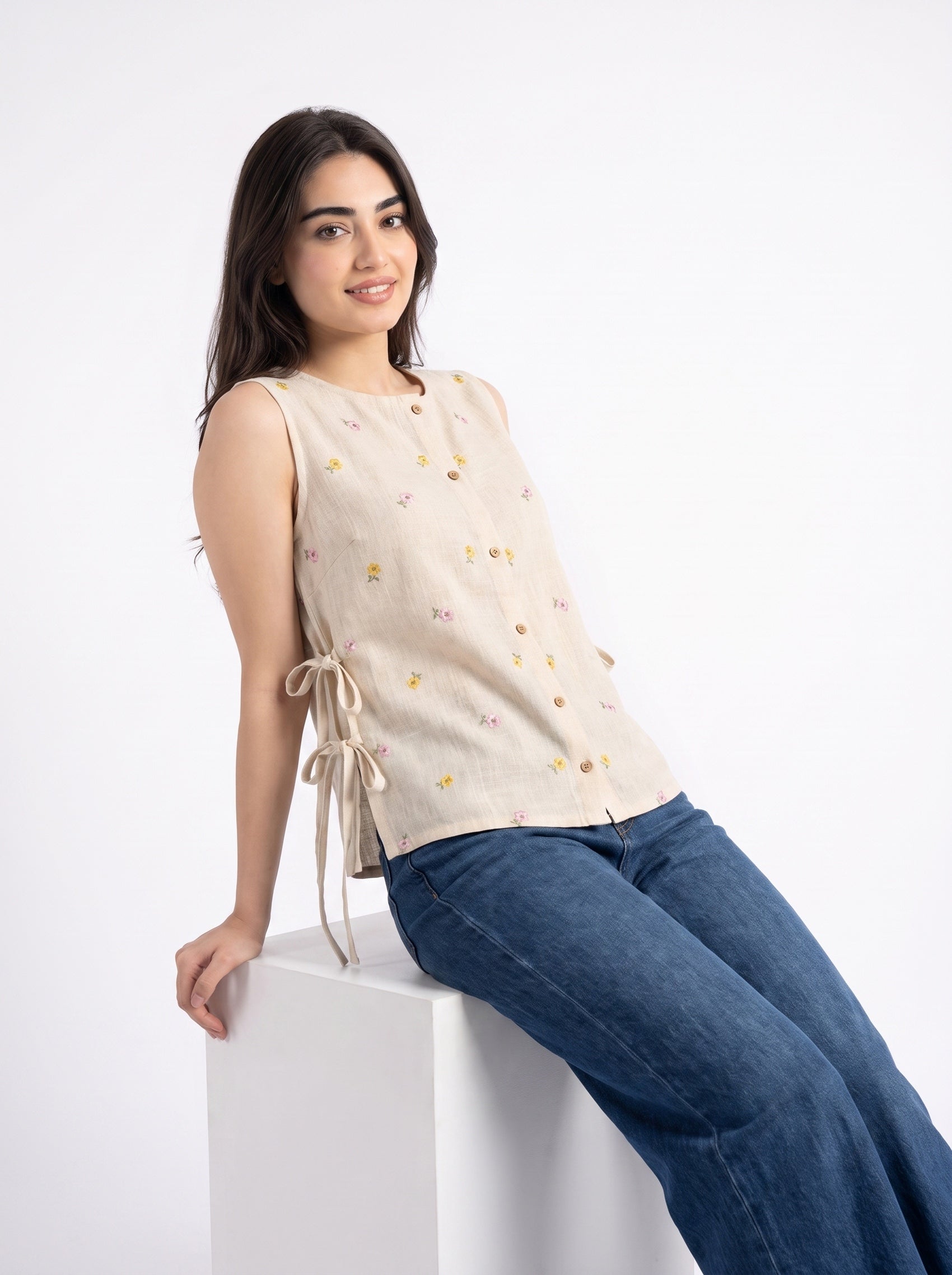 A woman wearing a sleeveless beige linen top with small embroidered floral details and side tie closures, paired with blue jeans, sitting on a white display block against a white studio background.