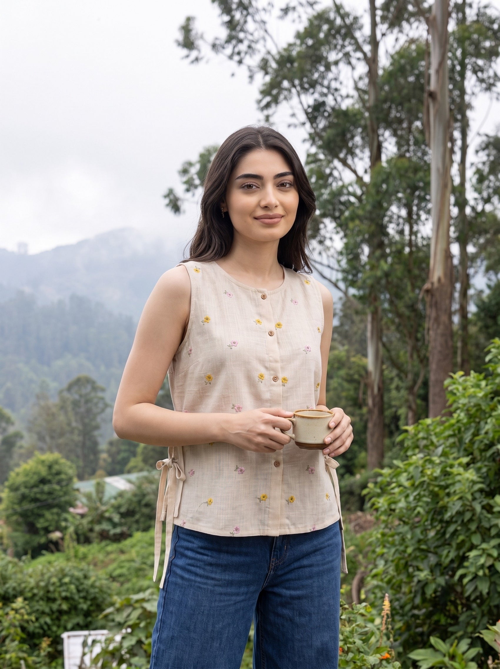 A woman wearing a sleeveless beige linen top with small yellow and pink floral prints, paired with blue jeans, holding a ceramic cup in an outdoor garden setting.