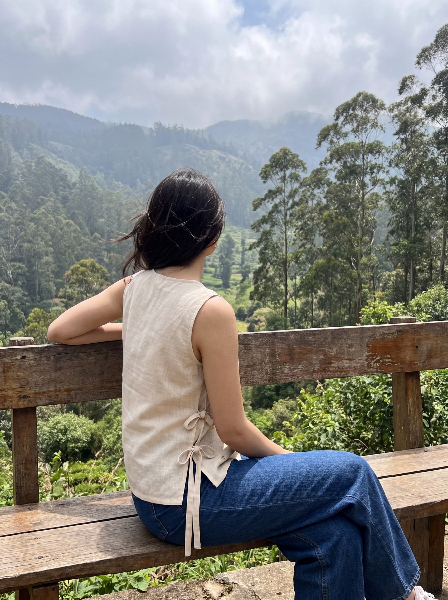 A woman sits on a wooden bench overlooking a lush green mountain valley, wearing a sleeveless beige linen top with side ties and blue jeans.