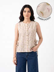 A woman modeling a sleeveless beige top with a subtle embroidered pattern, paired with blue jeans, against a plain white background. An inset circle in the top right highlights the embroidery detail with the text 'EMBROIDERY'.