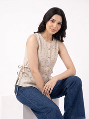 A woman wearing a sleeveless beige top with a subtle leaf pattern and blue jeans, sitting on a white block against a plain white background.