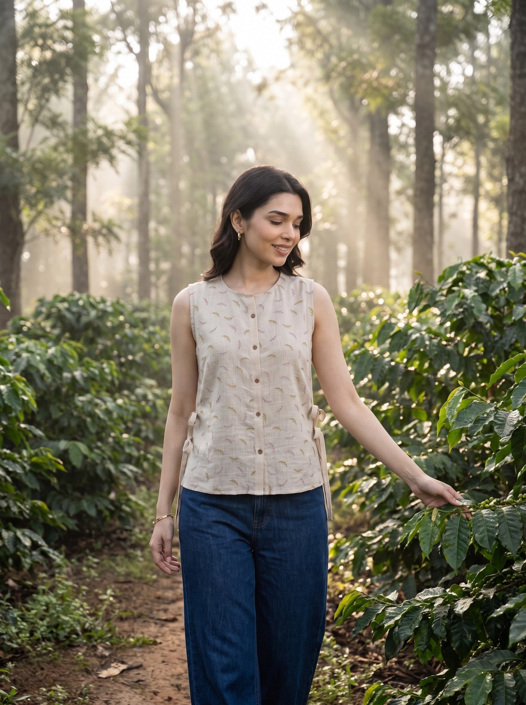 A woman stands in a misty forest of coffee plants, wearing a sleeveless beige top with a subtle leaf pattern and dark blue wide-leg pants.