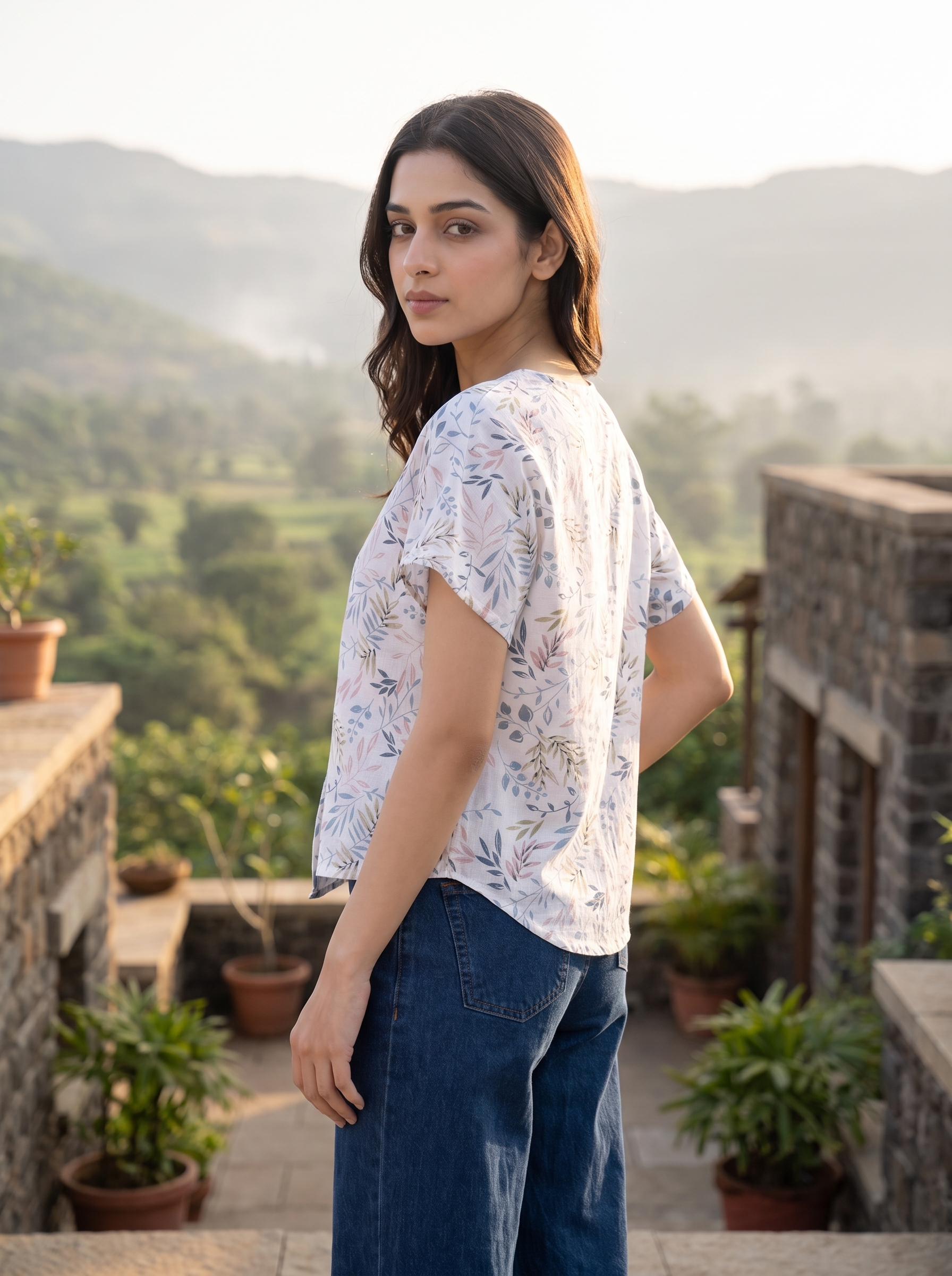 A woman wearing a white short-sleeve blouse with a delicate blue and green floral print, paired with dark blue jeans, walking along a mountain path with misty peaks in the background.