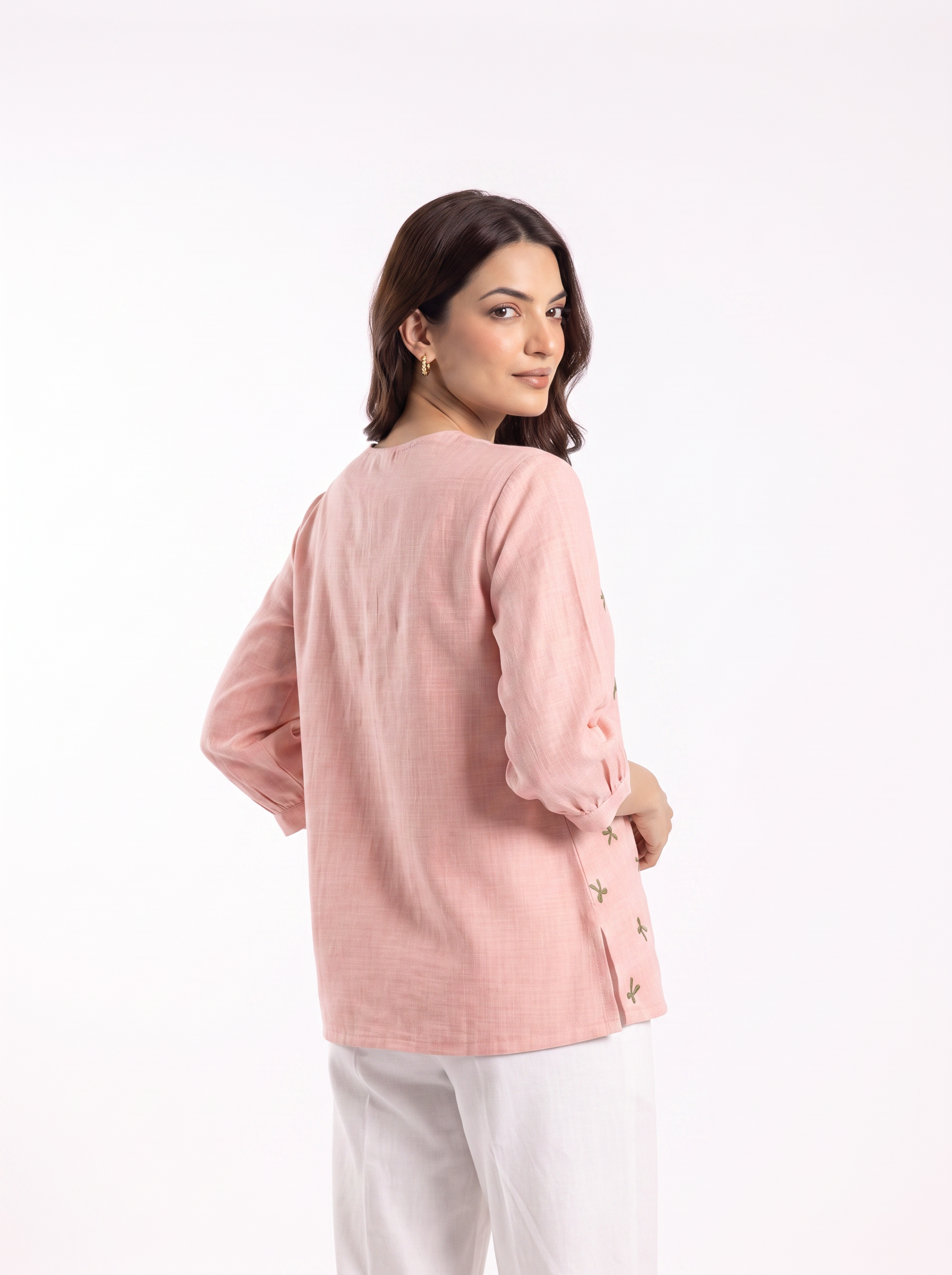 A woman wearing a light pink linen long-sleeve top and white trousers, photographed from the back against a plain white studio background.