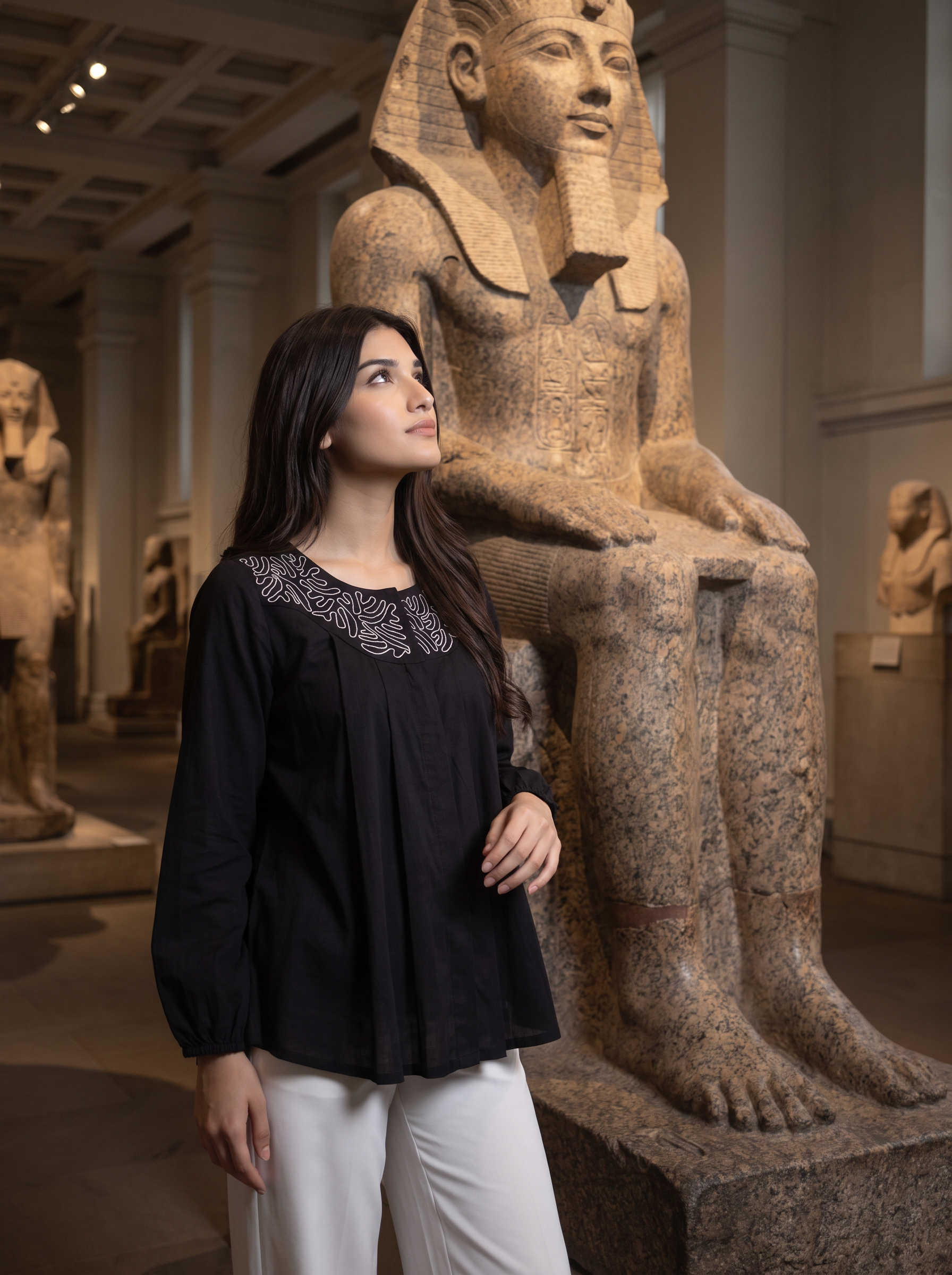 A woman wearing a black long-sleeve top with silver embroidery around the neckline and white pants, standing in a museum gallery next to a large stone pharaoh statue.