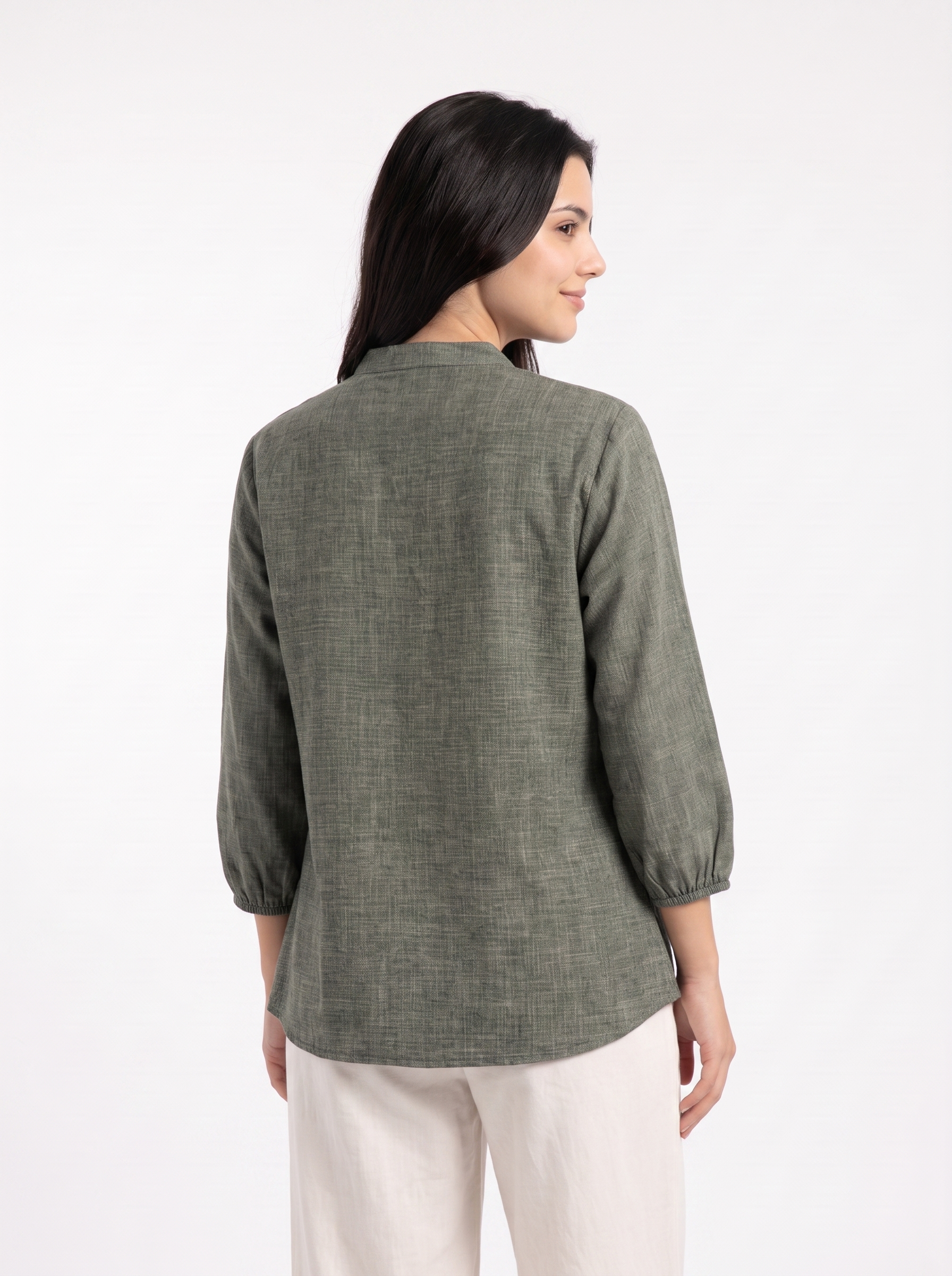 A woman shown from the back wearing a textured olive green long-sleeve top and off-white trousers, photographed against a clean white studio background.