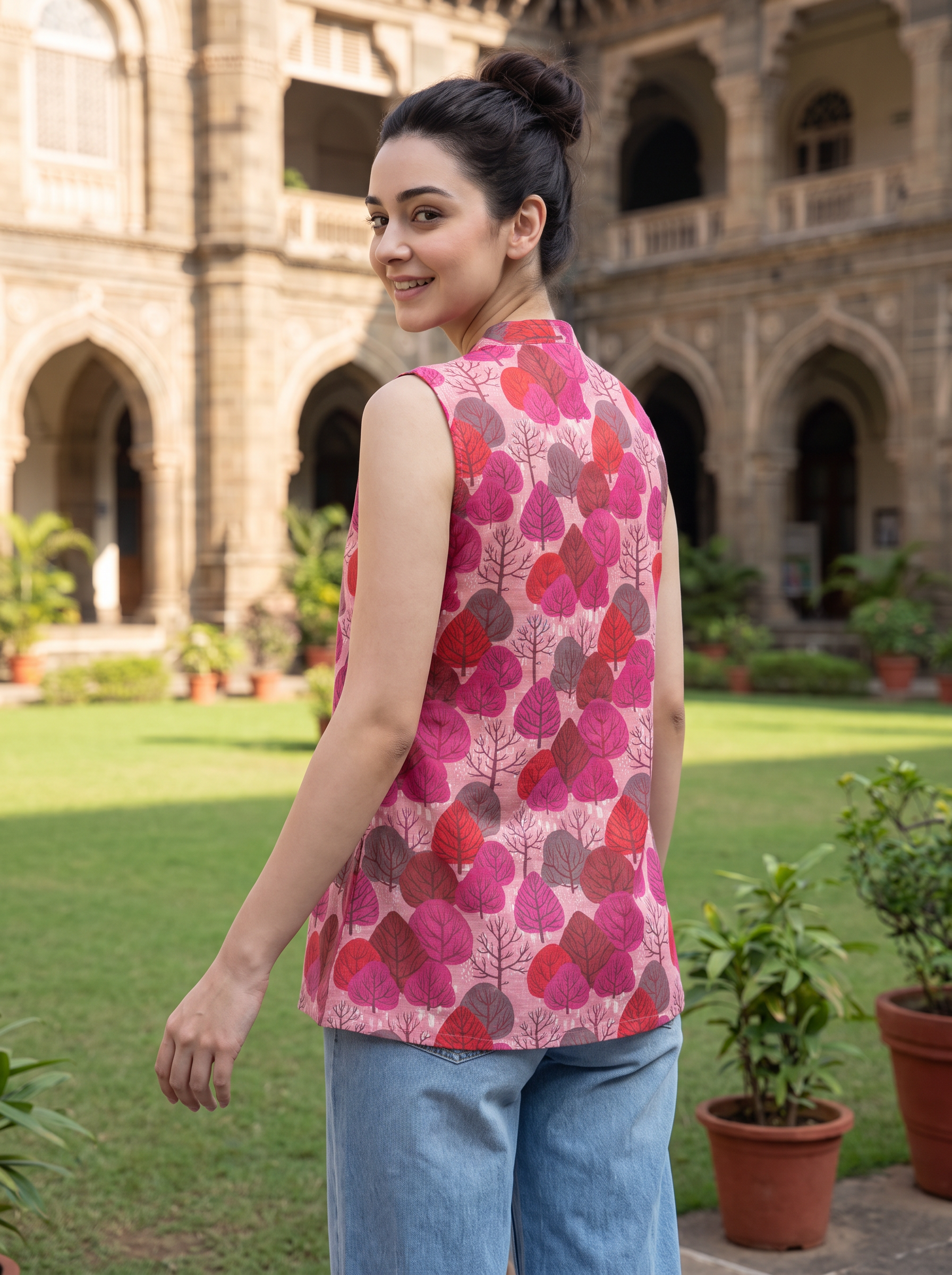A woman wearing a sleeveless pink vest with a red and magenta leaf pattern, paired with light blue jeans, standing in a garden courtyard with a historic stone building in the background.