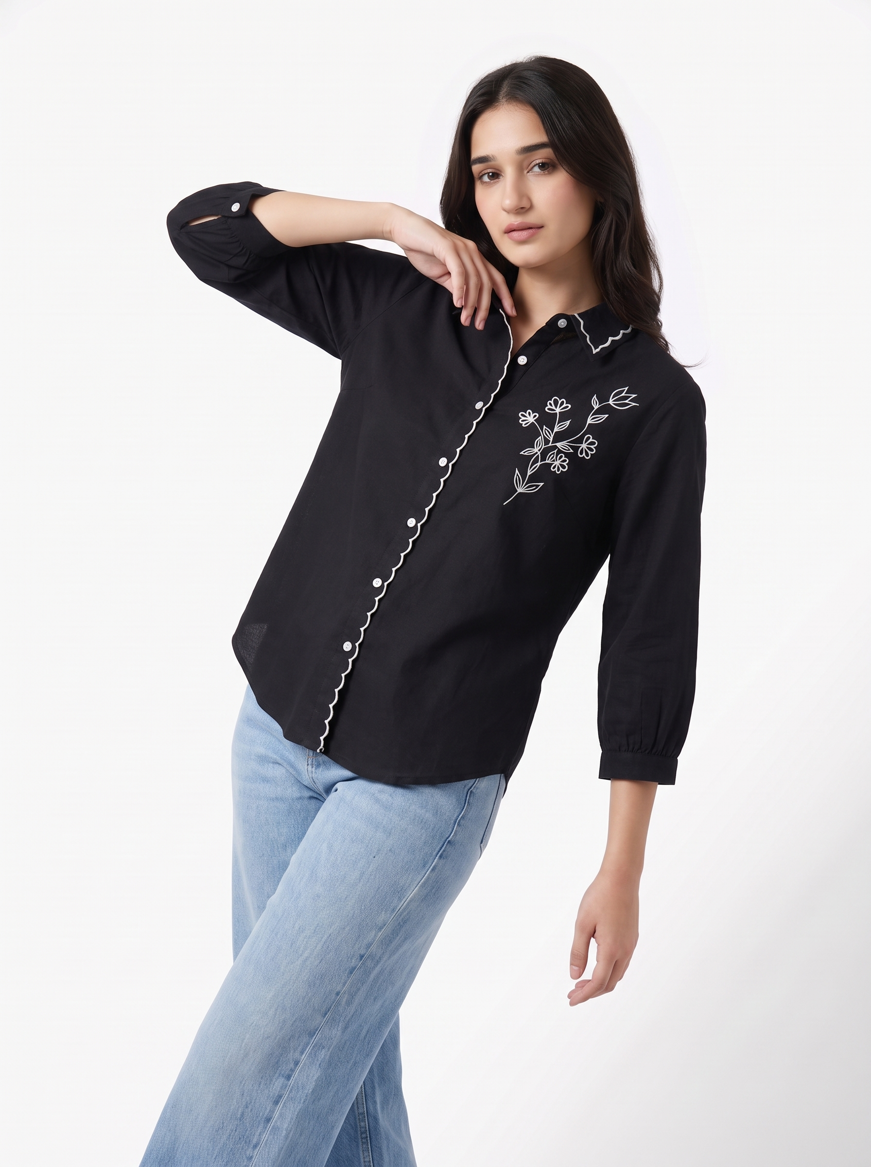 A woman modeling a black long-sleeve button-up shirt with white floral embroidery on the chest and a decorative white chain detail along the front placket, paired with light blue jeans against a plain white studio background.