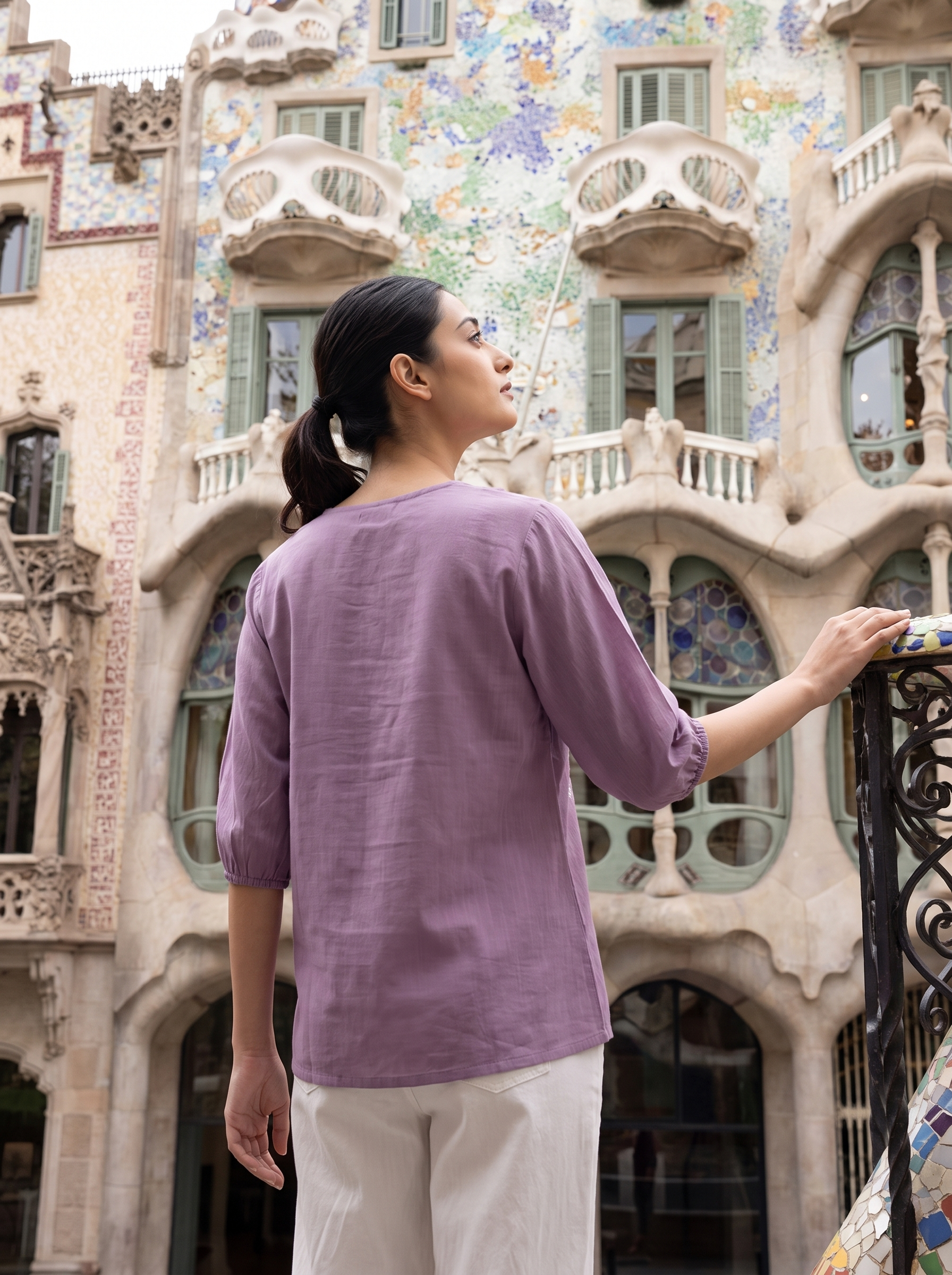 A woman wearing a lavender linen-blend top and white pants stands in front of the colorful Gaudí architecture of Casa Miléu in Barcelona, viewed from behind to show the back of the top.