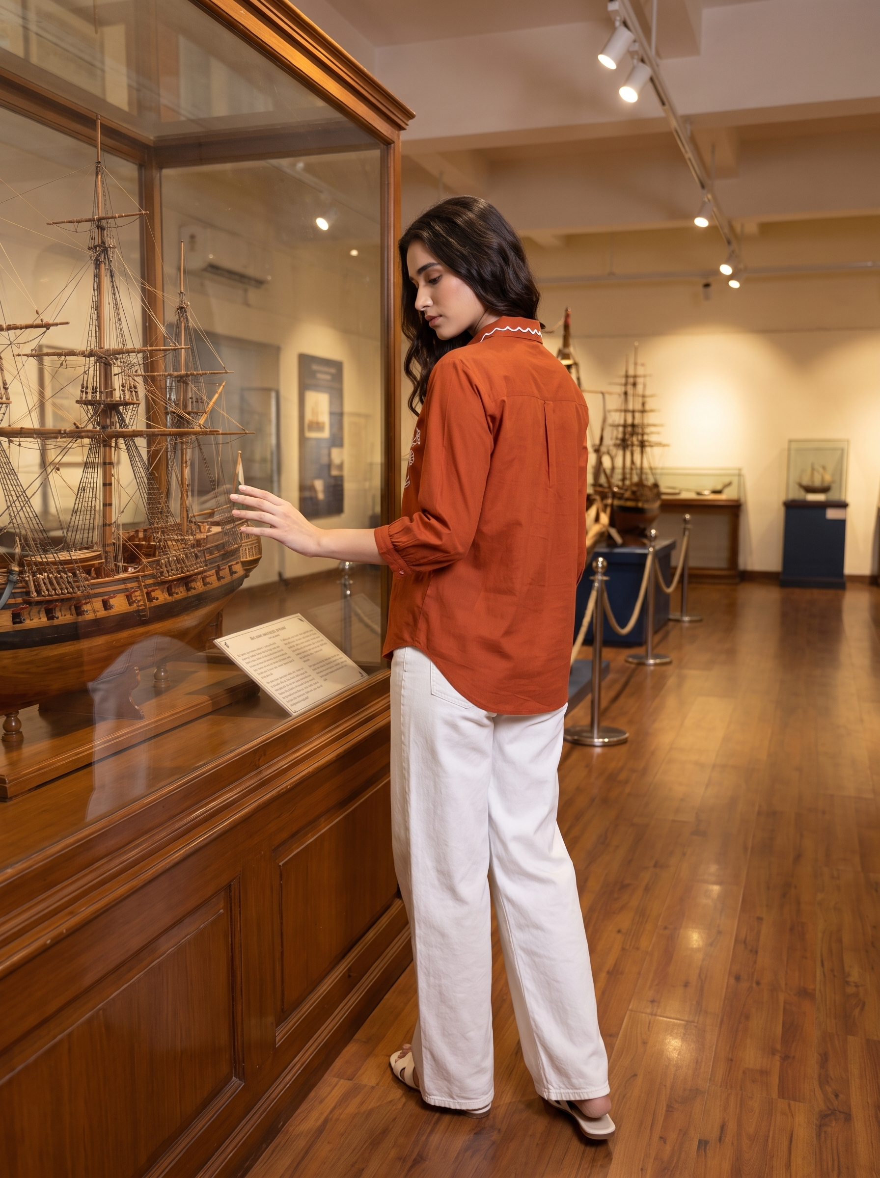 A woman in an orange-brown long-sleeve shirt and white wide-leg pants stands in a museum gallery, touching a large wooden ship model displayed inside a glass case.
