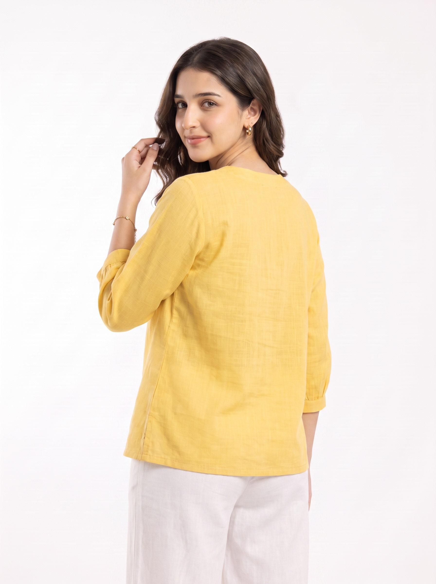 A woman wearing a yellow linen three-quarter sleeve top and white trousers, photographed from the back against a plain white studio background.