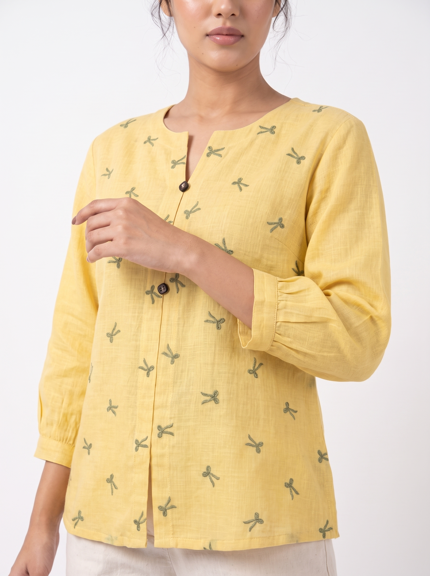 A woman wearing a yellow linen-style long-sleeve top with a split neckline, dark buttons, and a small green dragonfly print, photographed against a plain white studio background.