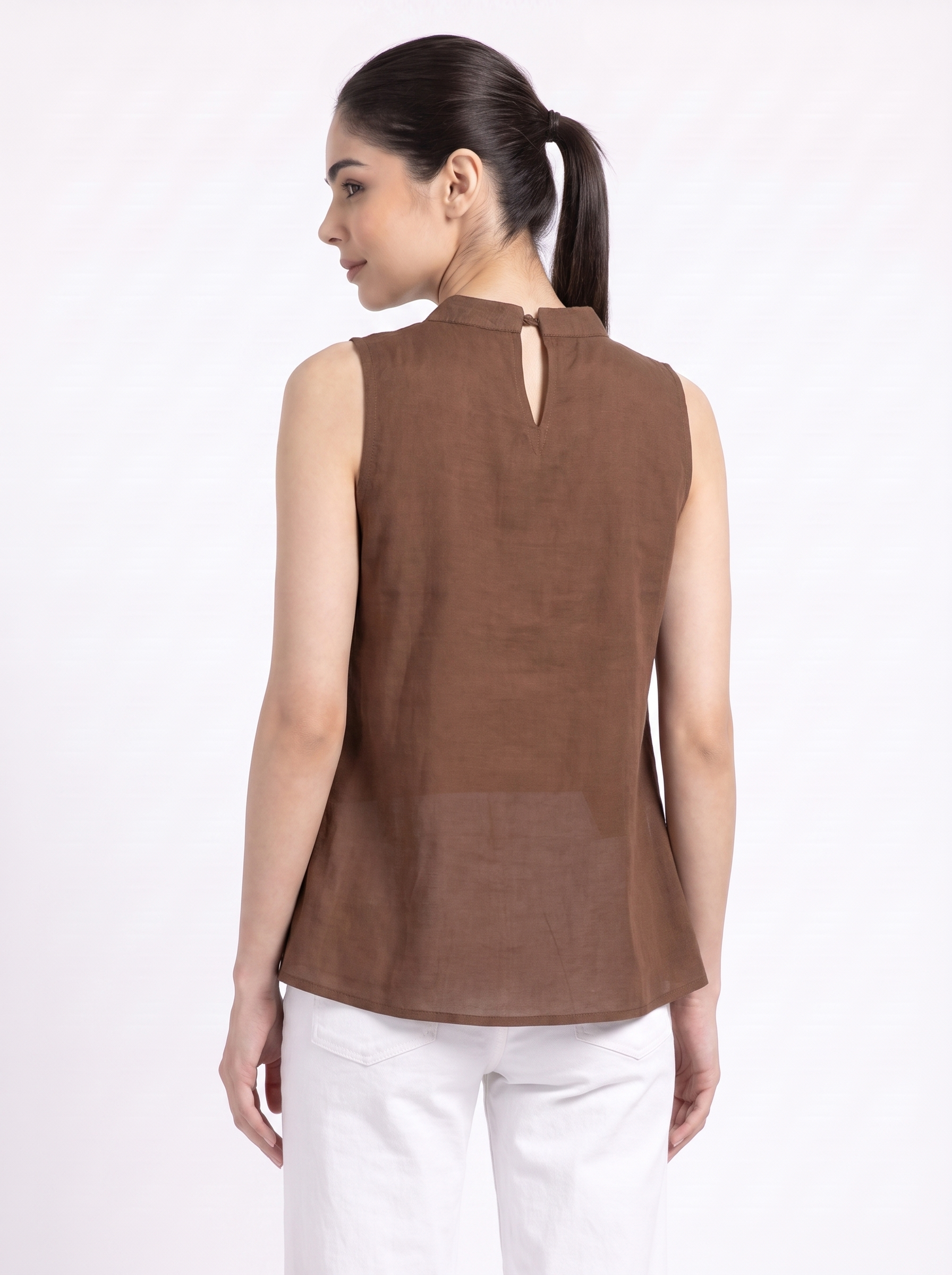 A woman shown from the back wearing a sleeveless brown top with a small keyhole cutout at the neckline, paired with white pants, against a plain white studio background.