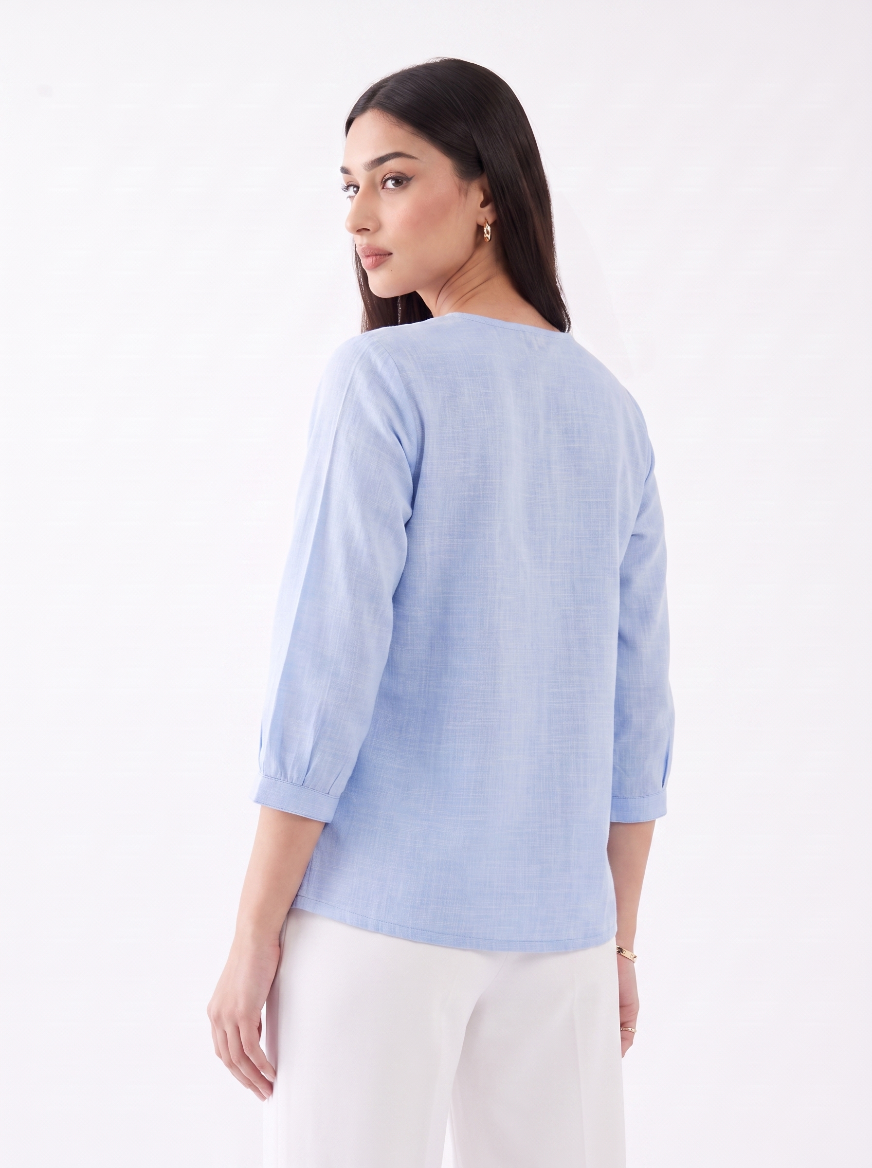 A woman shown from the back wearing a light blue three-quarter sleeve top and white pants, photographed against a plain white studio background.